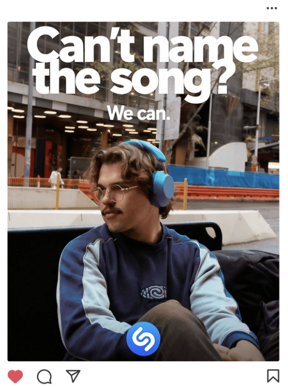 A young man with brown curly hair, glasses, and a mustache wearing large blue headphones, sitting outdoors on a bench with a black backpack beside him, in an urban setting with buildings and trees in the background, and a promotional text overlay asking if you can't name the song, with a Shazam logo on his sleeve.