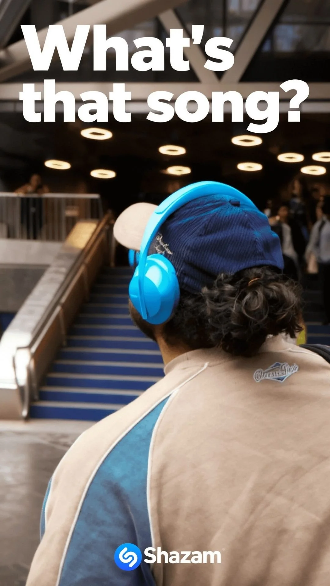 Back view of a person with curly hair wearing a beige and blue jacket, blue corduroy hat, and bright blue headphones in a public outdoor space, with a staircase and other people in the background.