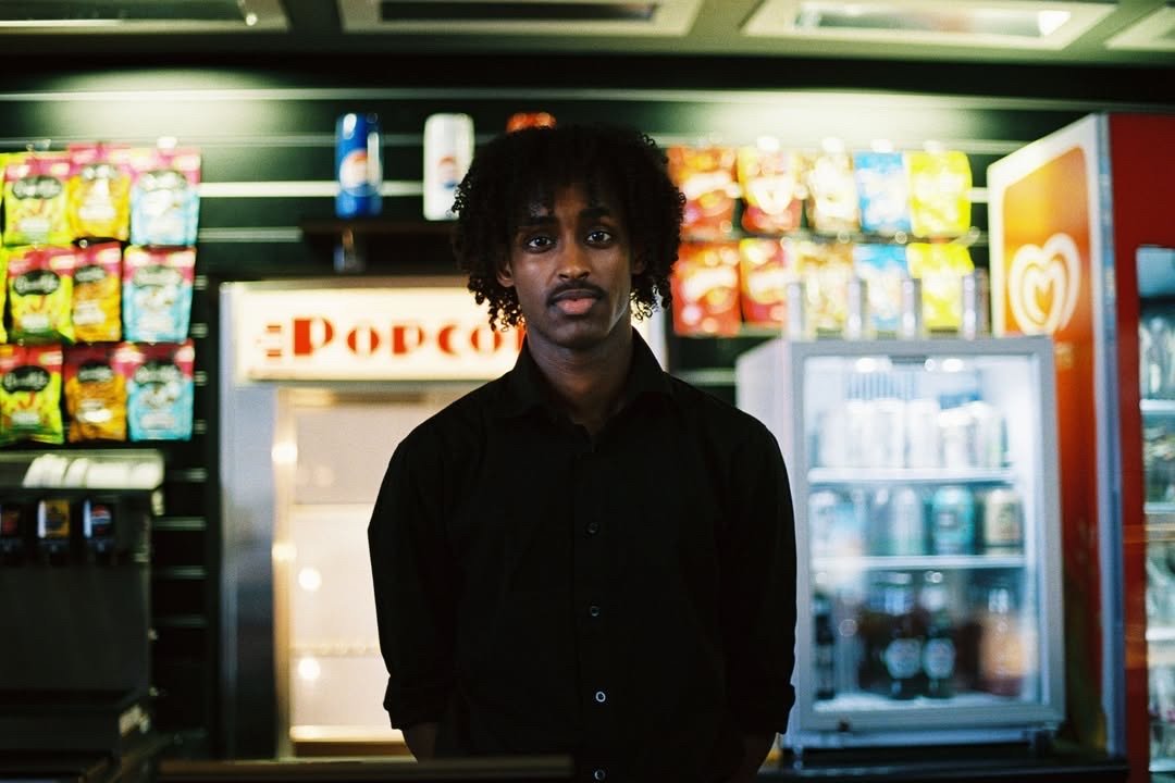 A man with curly hair and a black shirt standing in a convenience store, with a snack shelf and a frozen treats freezer in the background.
