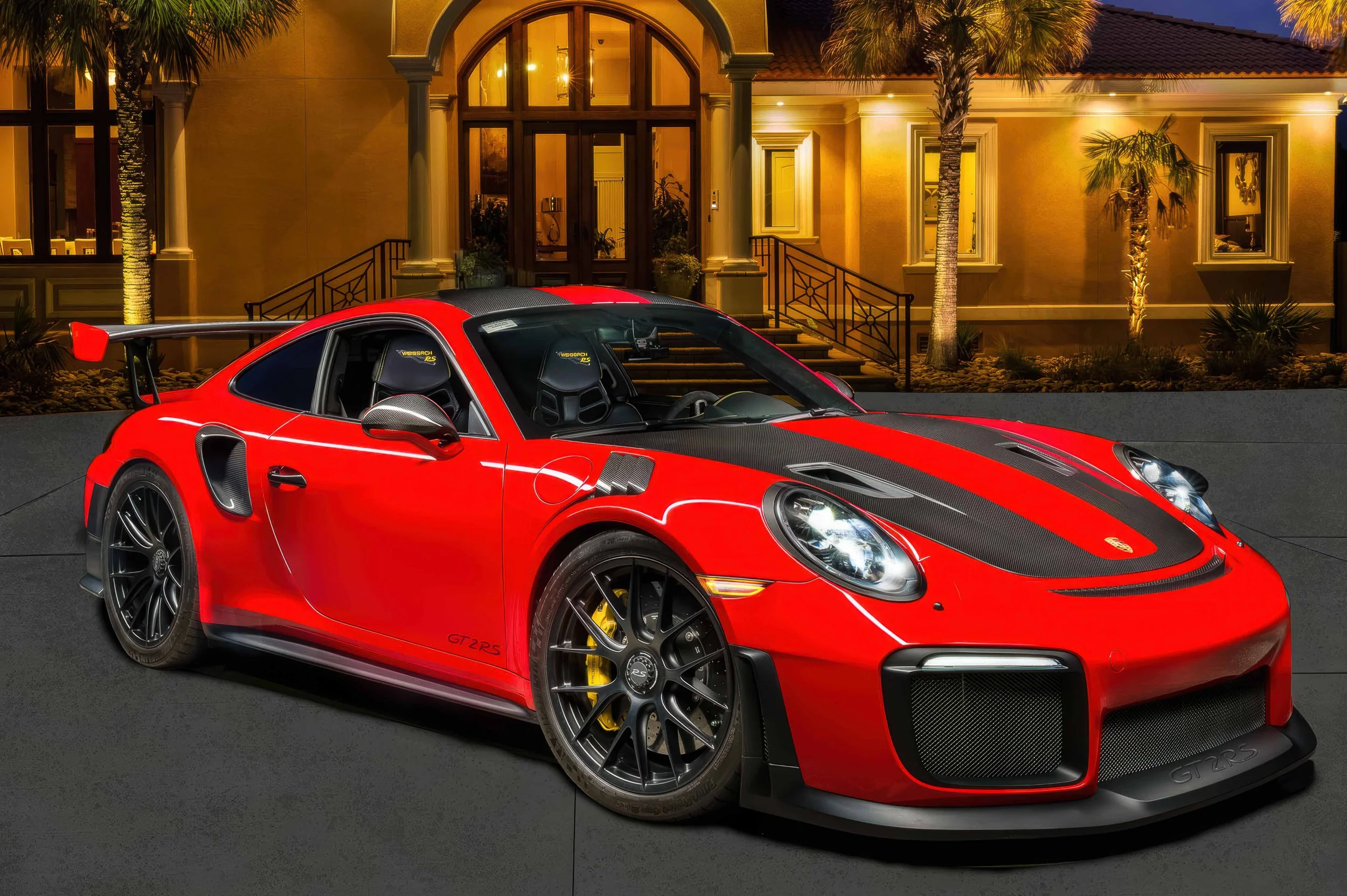 Red Porsche 911 GT2 RS photographed at dusk in front of an illuminated modern home, showcasing dramatic lighting and reflections.