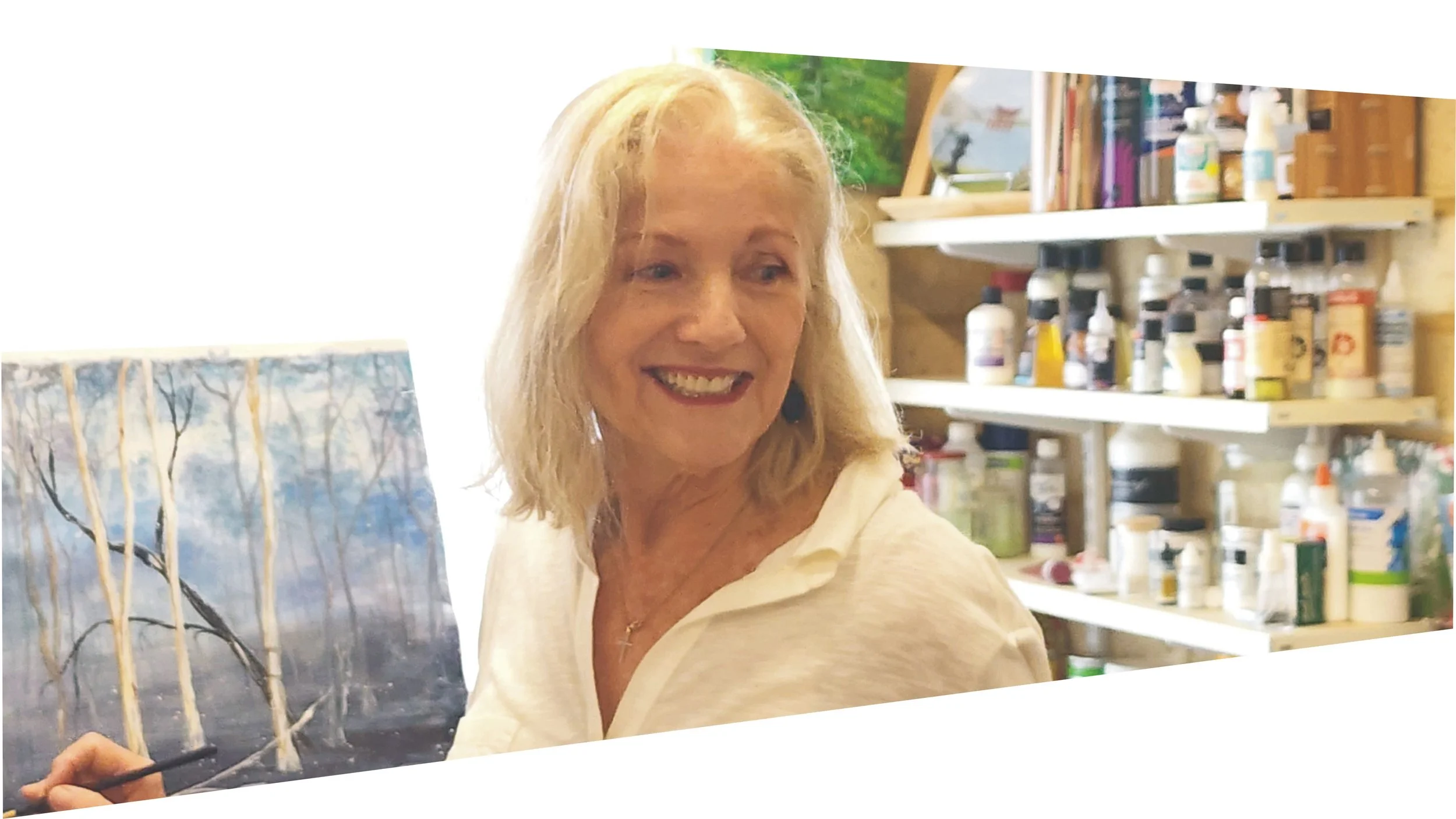 Artist Lauree in her happy space,  in her art studio in Helena Valley, Perth Western Australia.  Surrounded by shelves of paints and art supplies and working on a landscape painting with Ghost Gum trees.