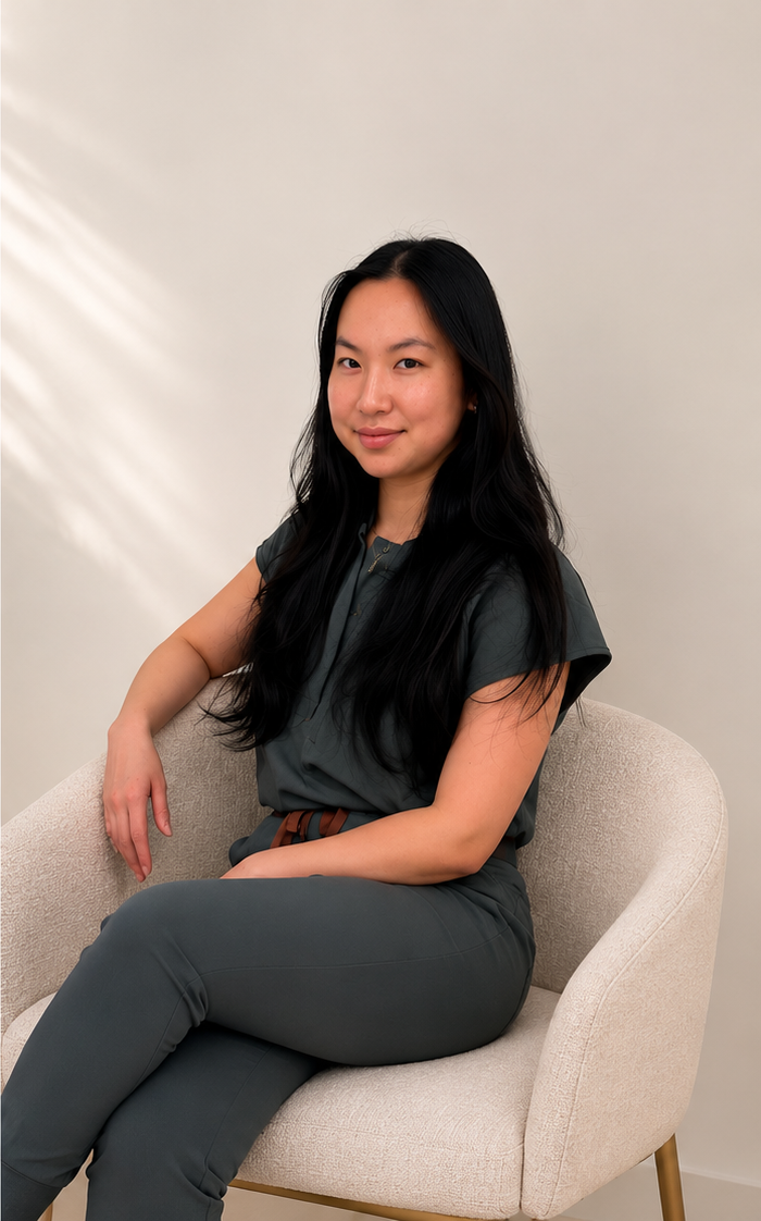 Woman sitting on beige armchair against plain wall, wearing dark gray outfit with brown belt, long black hair, smiling, relaxed posture