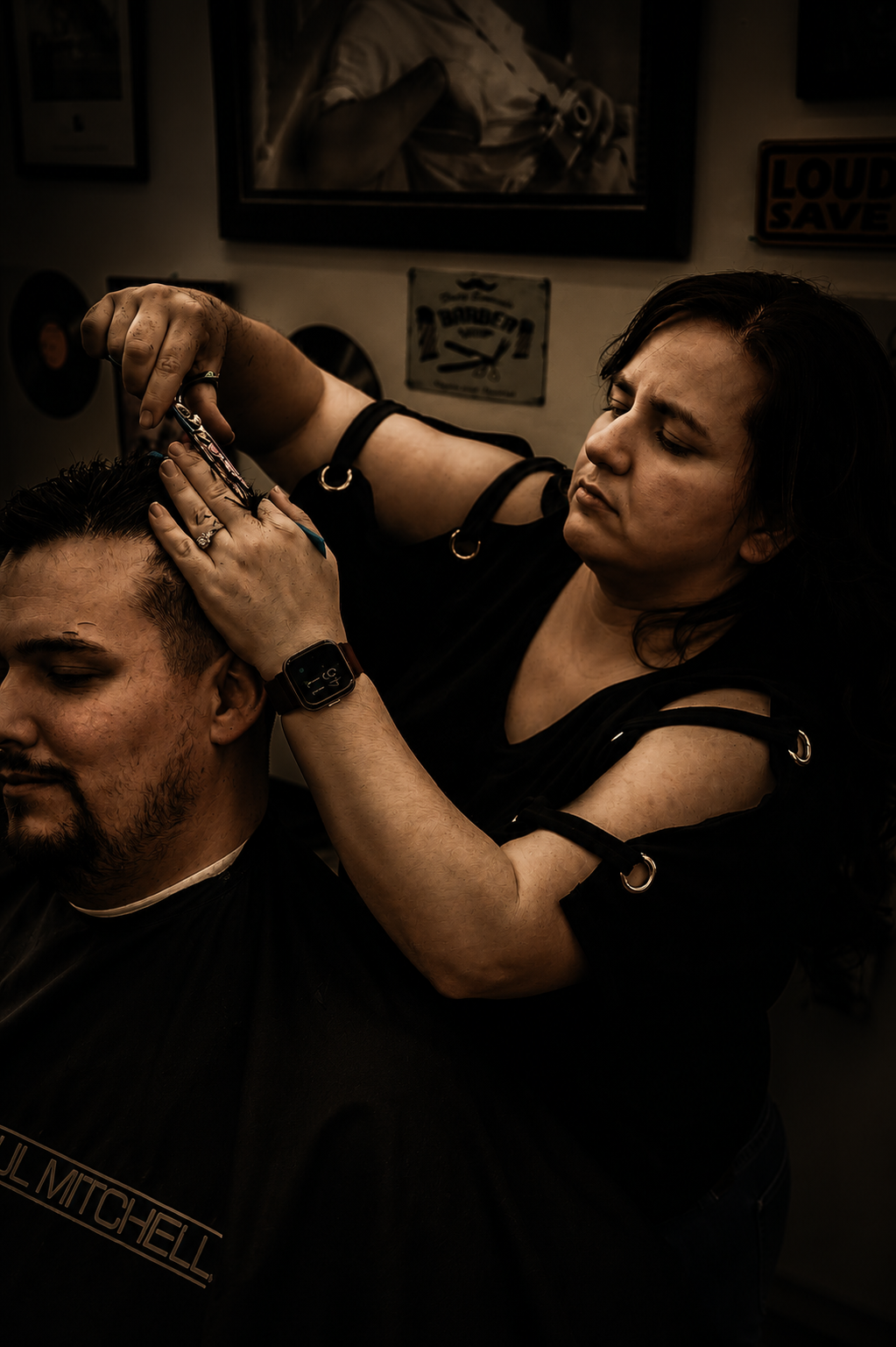 A female barber cutting a male client's hair in a barbershop, with framed pictures and signs on the wall in the background.