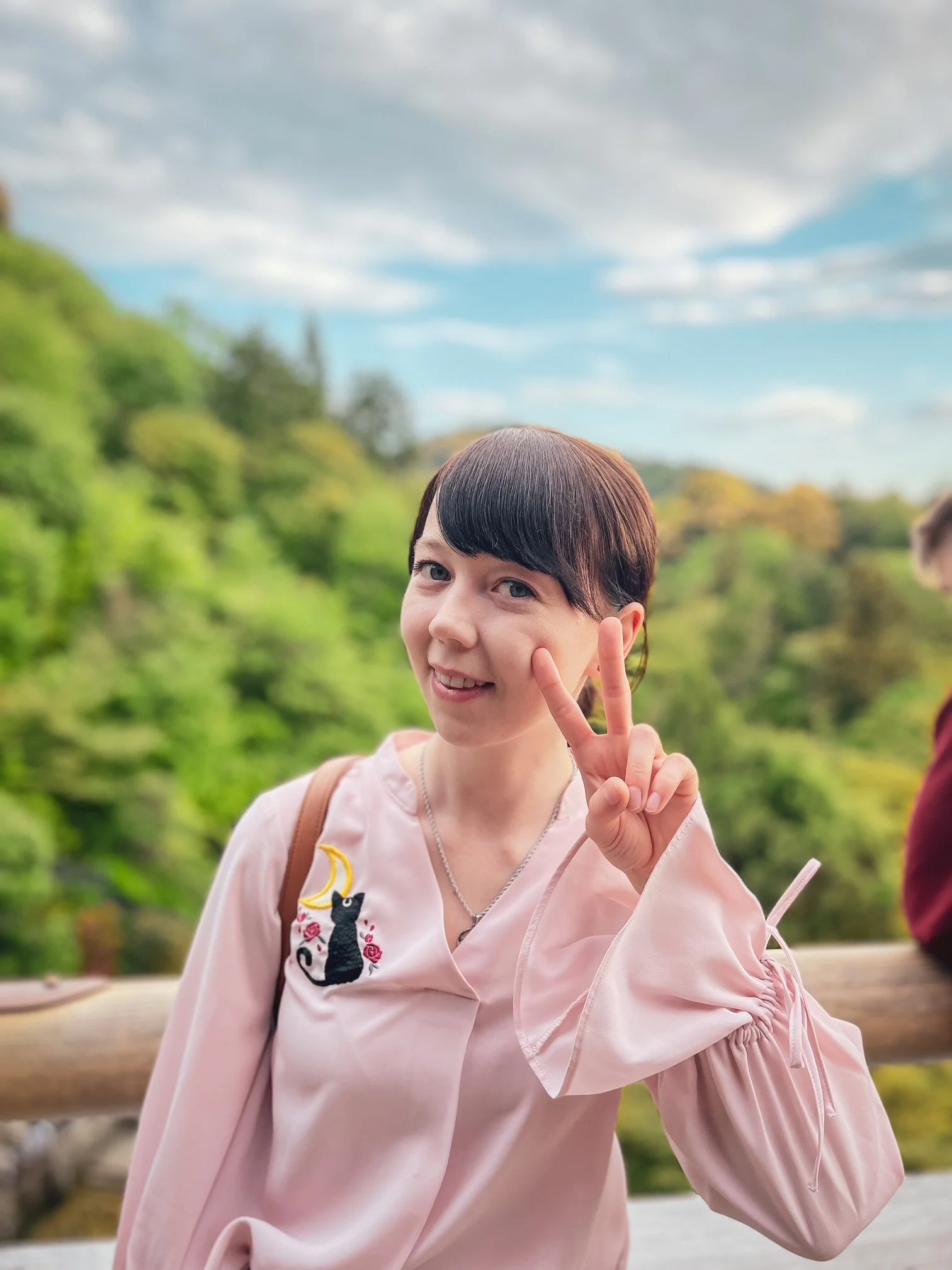 A girl with short dark hair smiling and making a peace sign with her right hand outdoors. She is wearing a light pink jacket with a black cat embroidery and a necklace. The background has green trees and a cloudy sky.