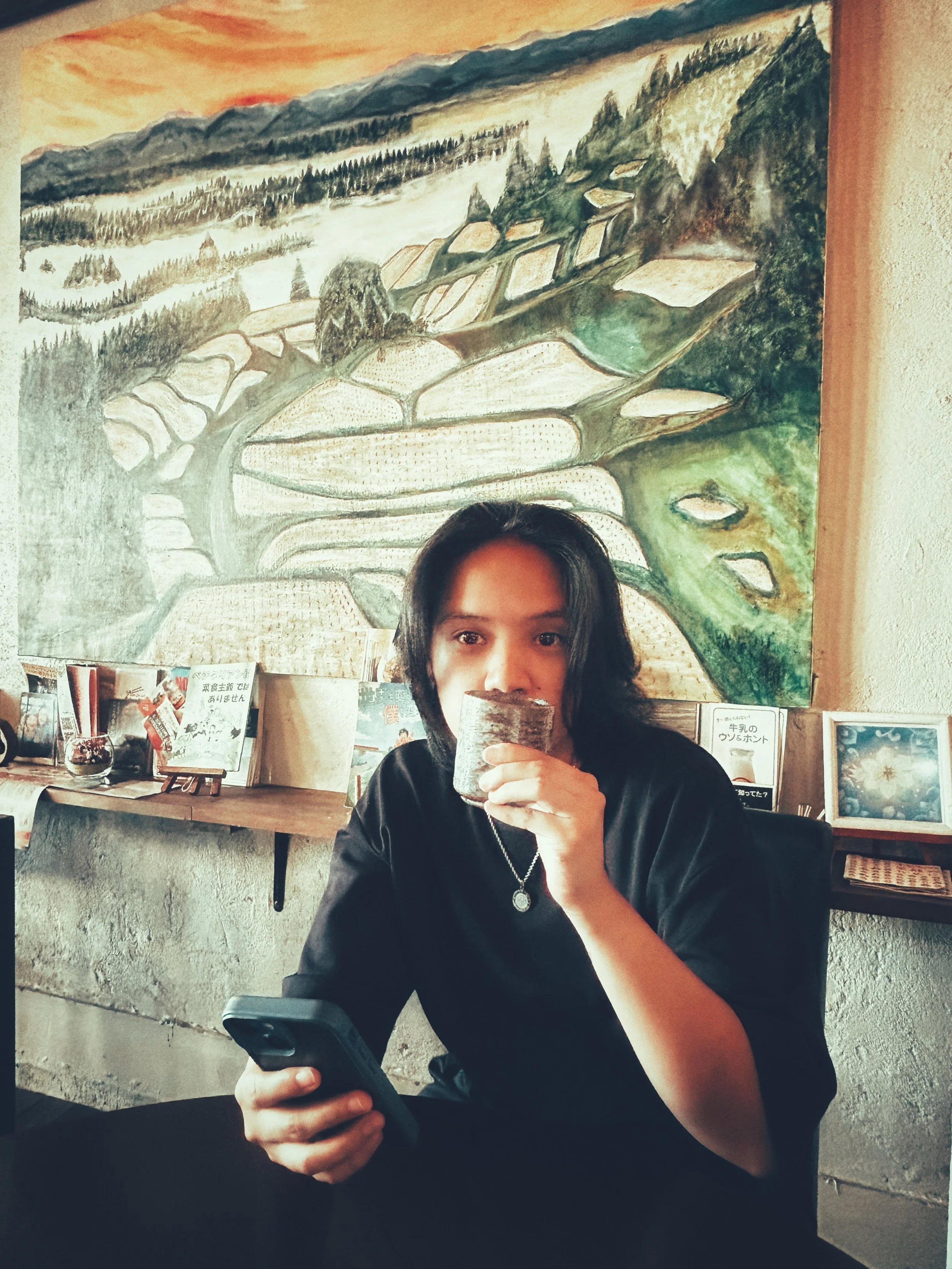 A person with dark hair sitting at a table in a restaurant, holding a mobile phone in one hand and a beverage wrapped in a napkin in the other hand, with a large landscape painting of terraced fields and mountains on the wall behind them.