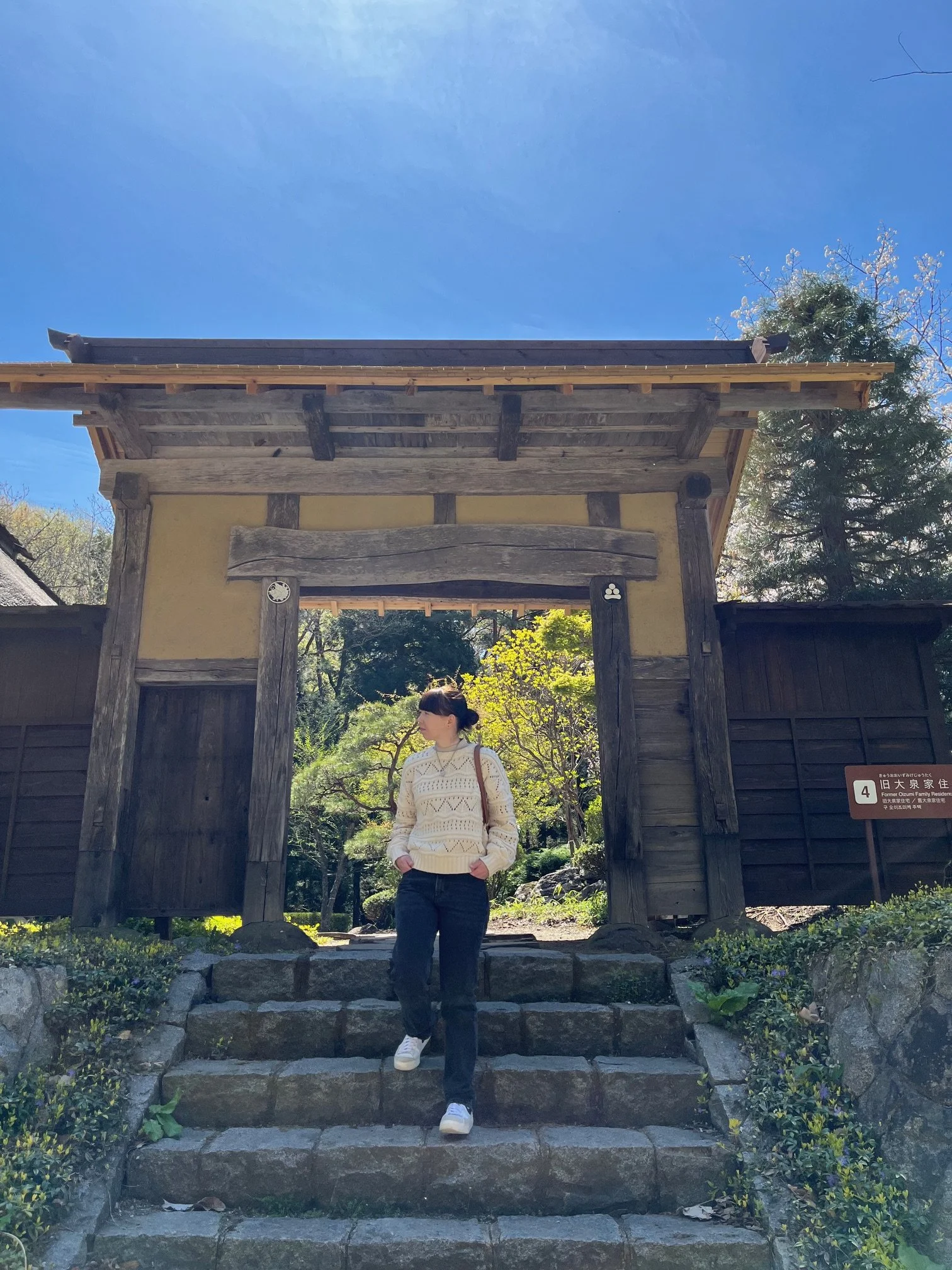 A woman standing on stone steps under a traditional wooden gate with a thatched roof, surrounded by trees and greenery, on a sunny day.