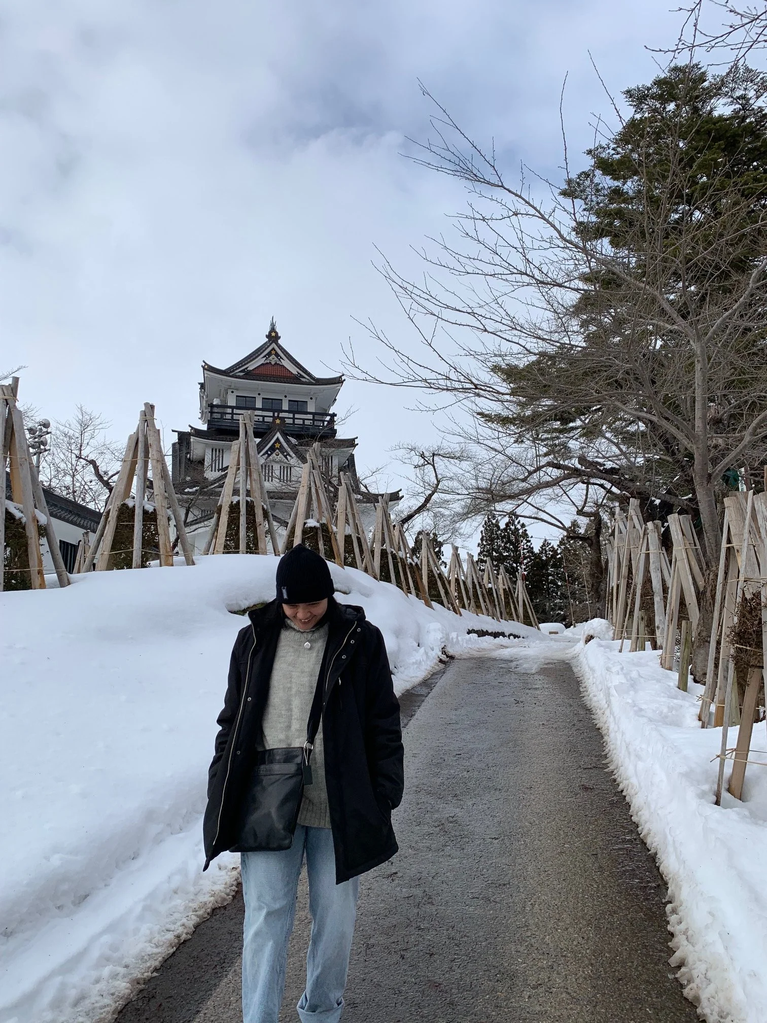 A woman walking on a snow-covered path surrounded by snowbanks, with a traditional Japanese castle in the background and leafless trees on either side.