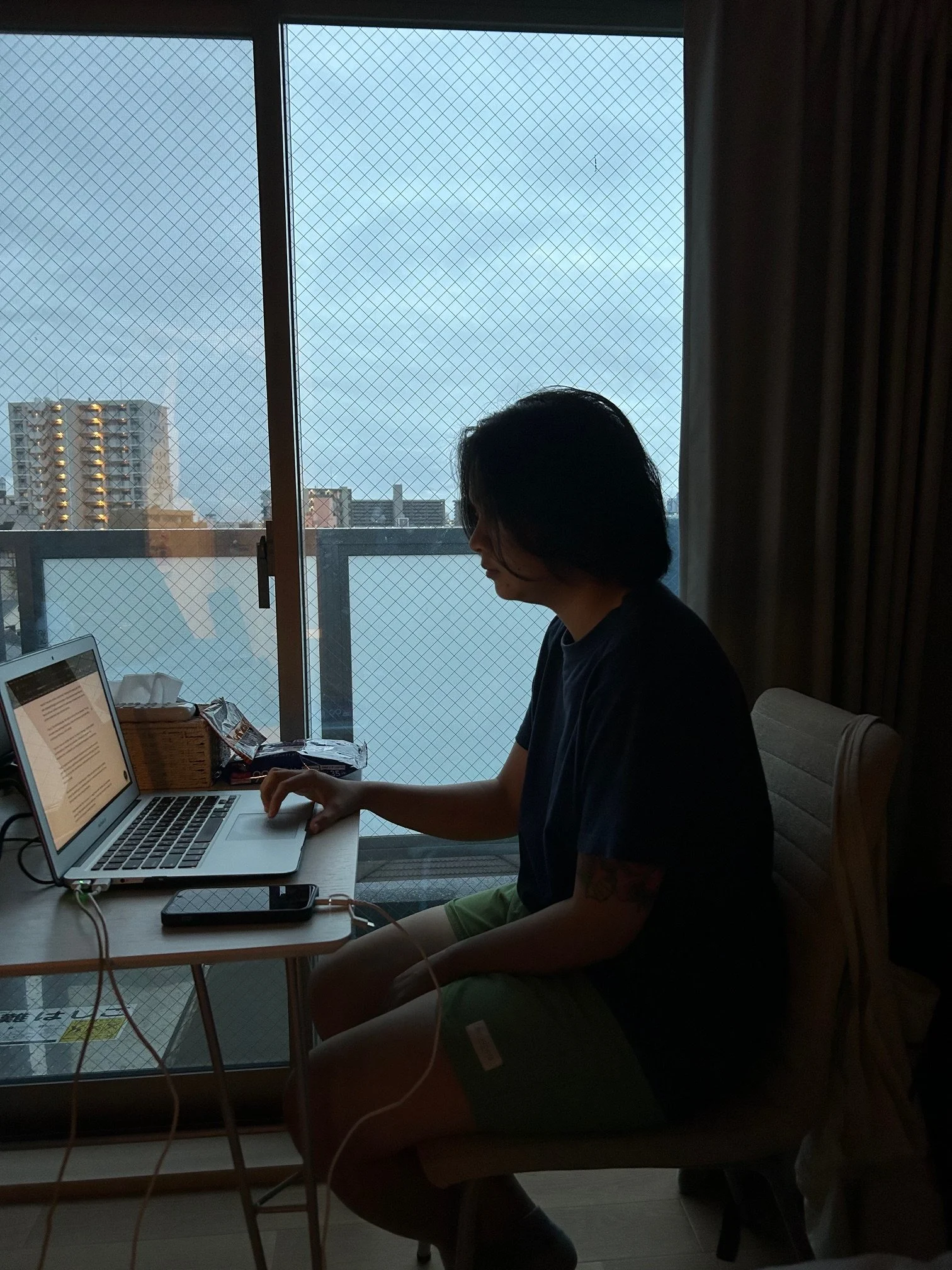 A person with black hair sitting at a table, using a laptop in a room with a sliding glass door and city buildings outside.