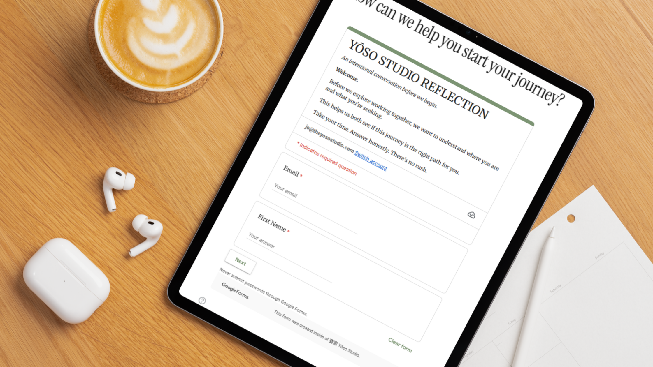 A wooden desk with a cup of coffee, a tablet displaying a reflection studio website, white wireless earbuds, a white wireless earbud case, and some white paper or notepad.