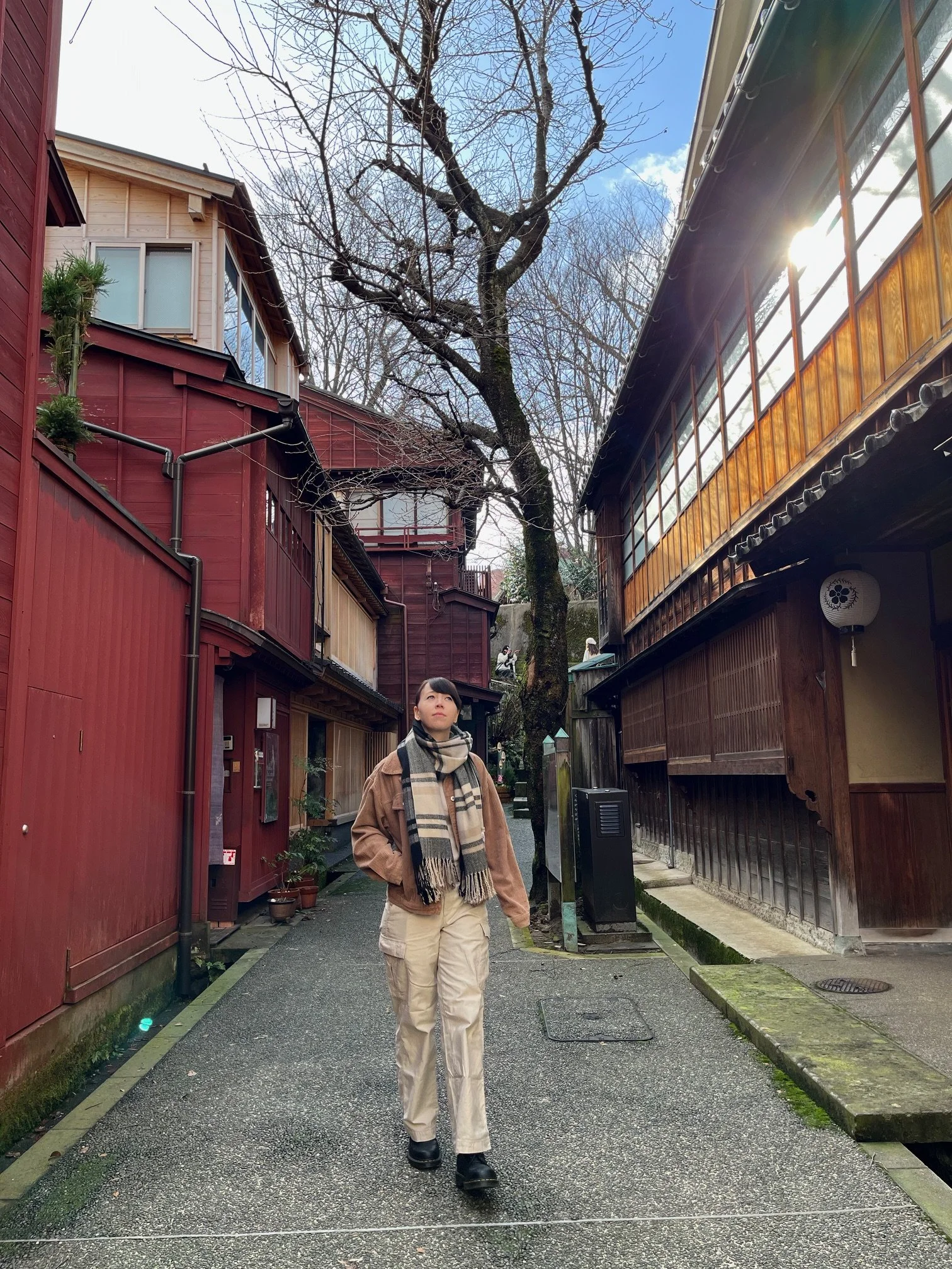 A person walking along a narrow alleyway between traditional Japanese buildings during daytime. The person is dressed in beige pants, a brown jacket, black shoes, and a plaid scarf. The alley has a tree with bare branches in the middle, and the sky is partly cloudy with sunlight coming from behind the building on the right.
