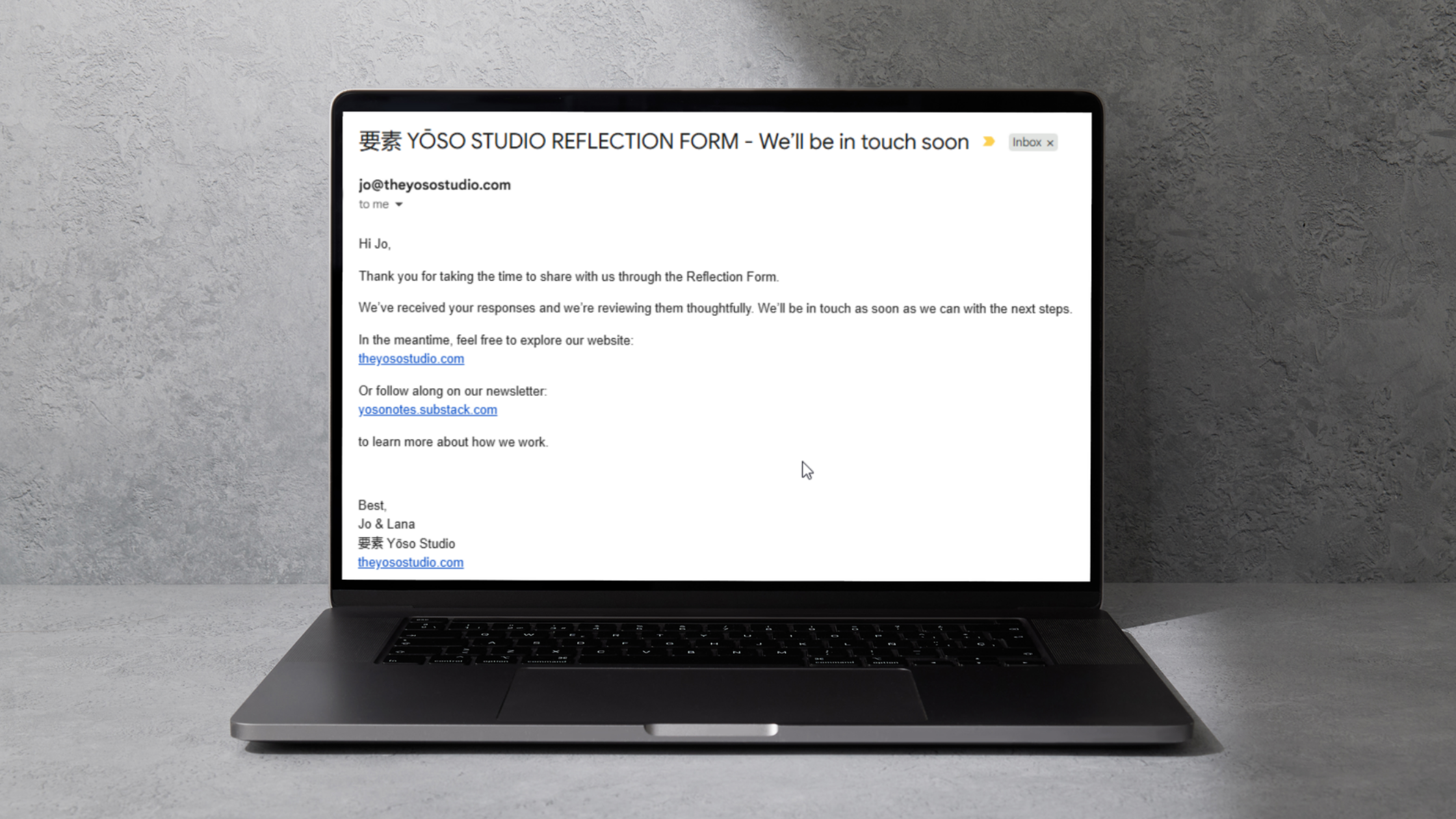 A laptop on a gray textured table displaying an email from Yōso Studio about a reflection form. The email contains text and links, with a gray concrete wall in the background.