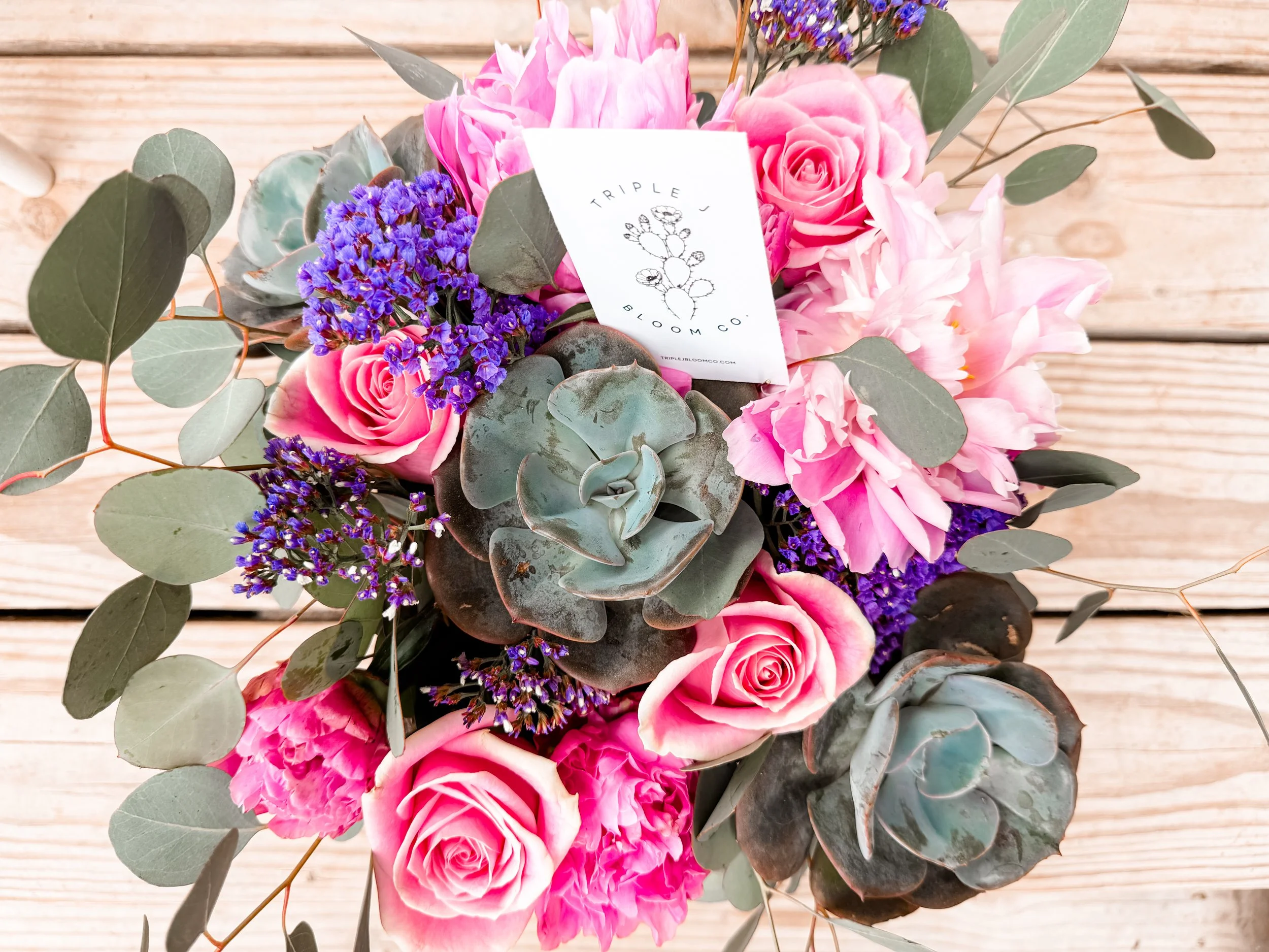 A bouquet of pink roses, purple statice, dusty miller, and eucalyptus leaves on a wooden surface, with a tag from Triple J Bloom Co.