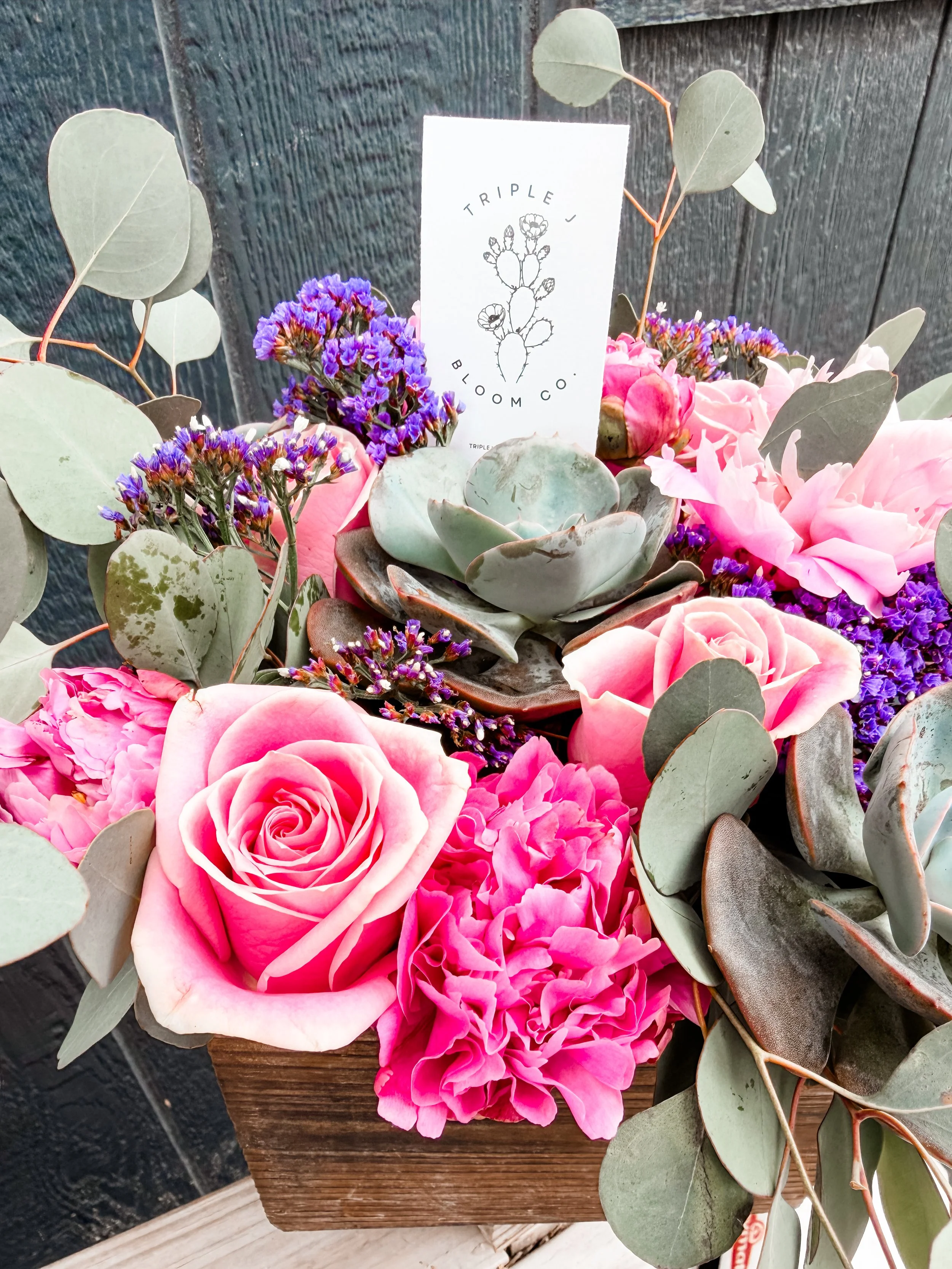 A floral arrangement featuring pink roses, purple statice, green eucalyptus leaves, and a white card with a black cactus design and the words 'Triple J Bloom Co.'
