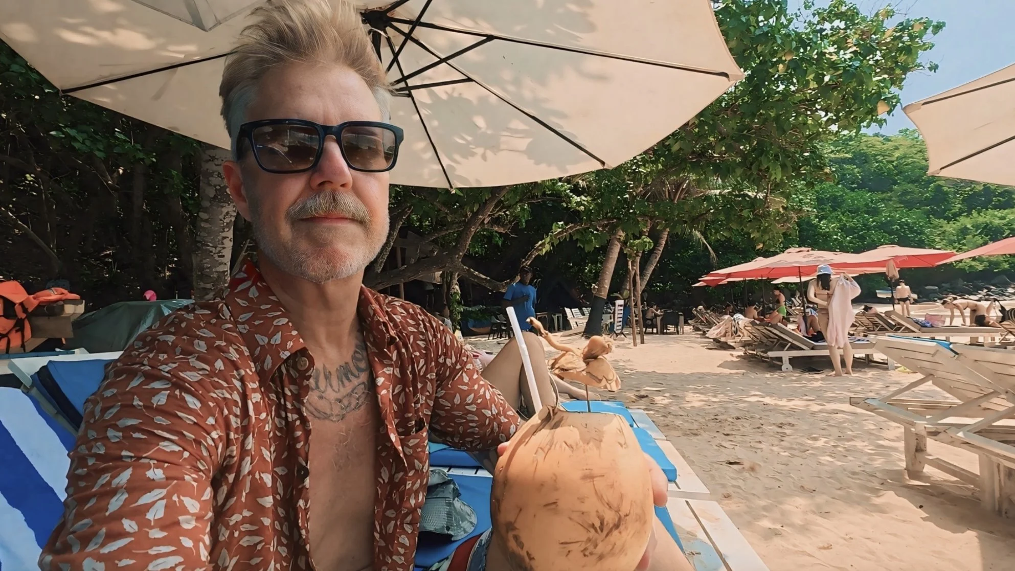 A man with sunglasses, a grey beard, and wearing a patterned shirt, sitting on a beach chair with a coconut drink, at a beach with umbrellas, lounge chairs, and people in the background.