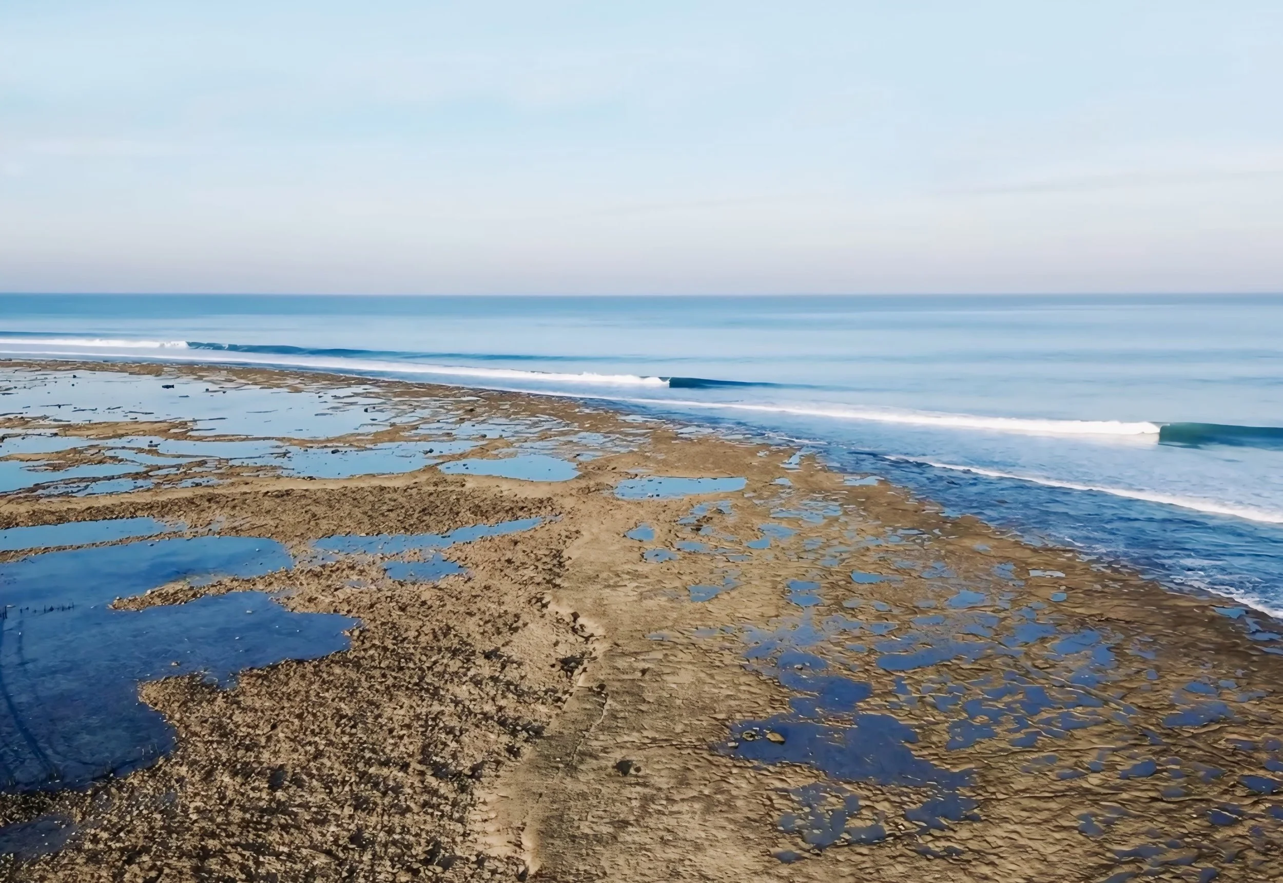 A sandy beach with tide pools and waves rolling in under a clear, blue sky.
