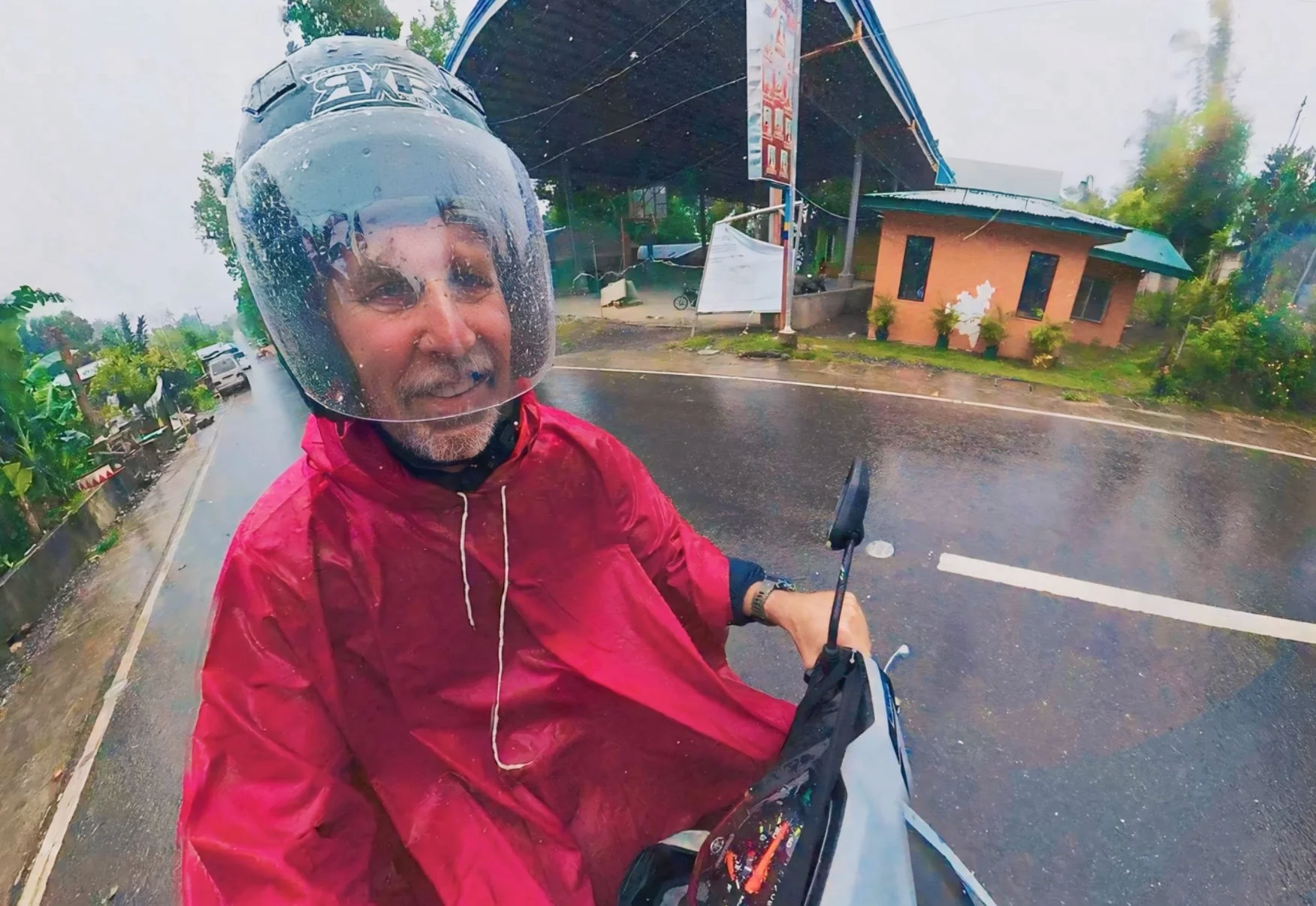 A man smiling while riding a motorcycle in the rain, wearing a clear helmet and a red rain jacket, on a wet street with buildings and trees in the background.