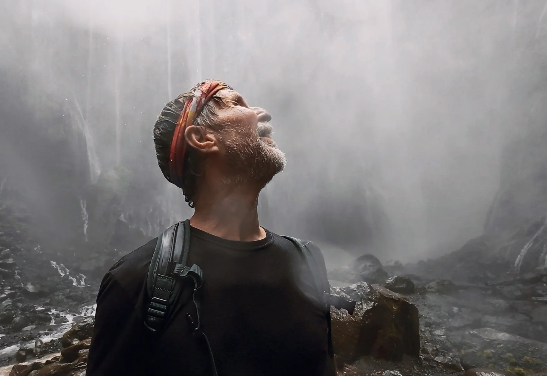 A man with gray hair and a beard wearing a black shirt and backpack stands in front of a large waterfall, with water cascading down behind him.