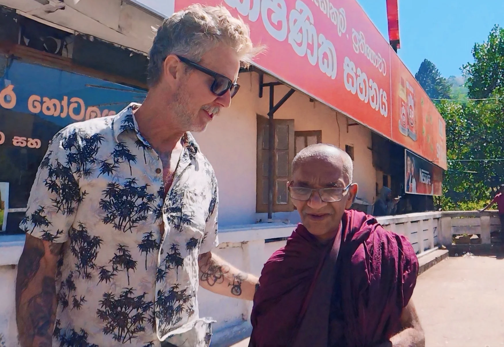 Two men are standing outside a building with blue and pink signage. One man has blonde hair, glasses, and is wearing a Hawaiian shirt. The other man is bald, wearing glasses, and dressed in maroon. They are smiling and appear to be engaged in a friendly interaction.