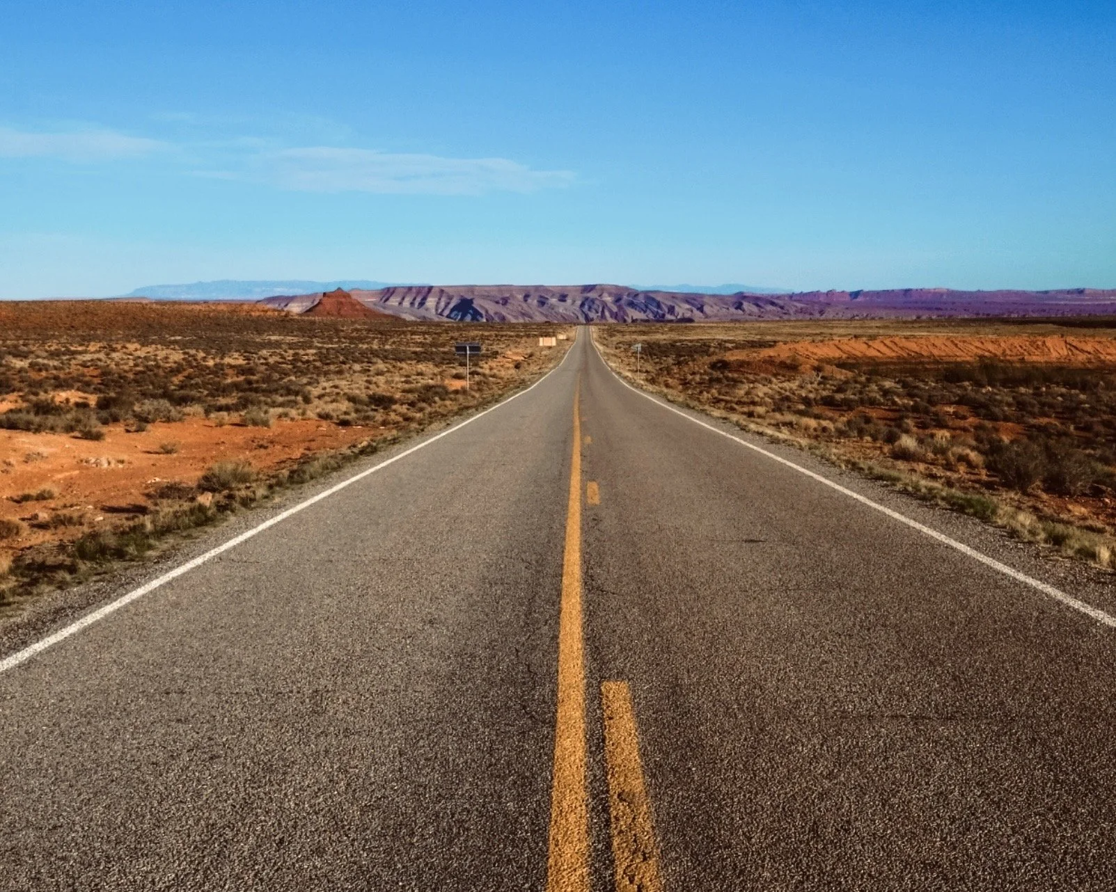 Long straight road stretching into the horizon in a desert landscape with colorful mesas under a blue sky.