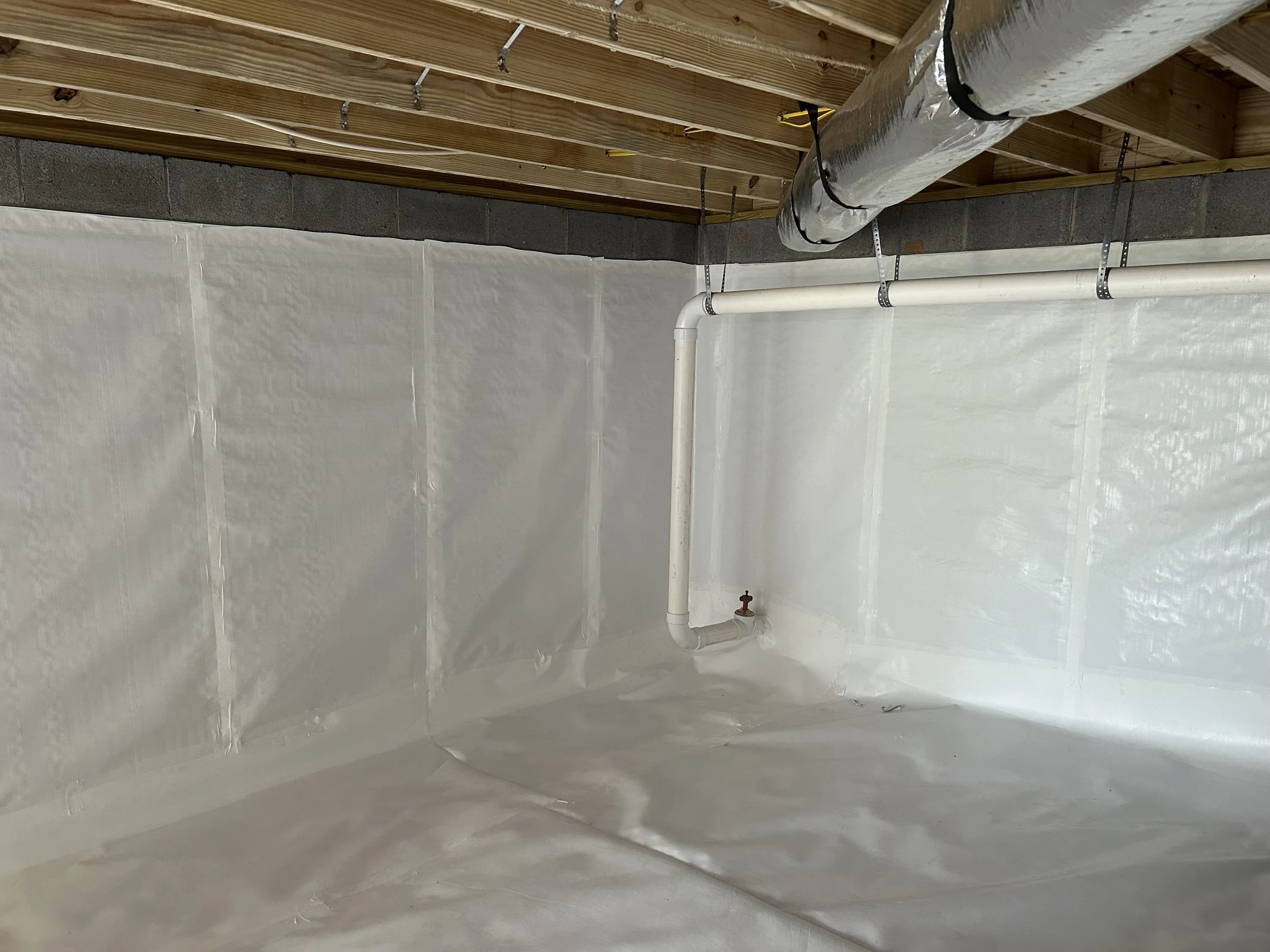A basement under construction with exposed wooden ceiling beams, a ventilation duct wrapped in silver insulation, and white plastic sheeting covering the walls, with a white pipe running vertically along the wall.