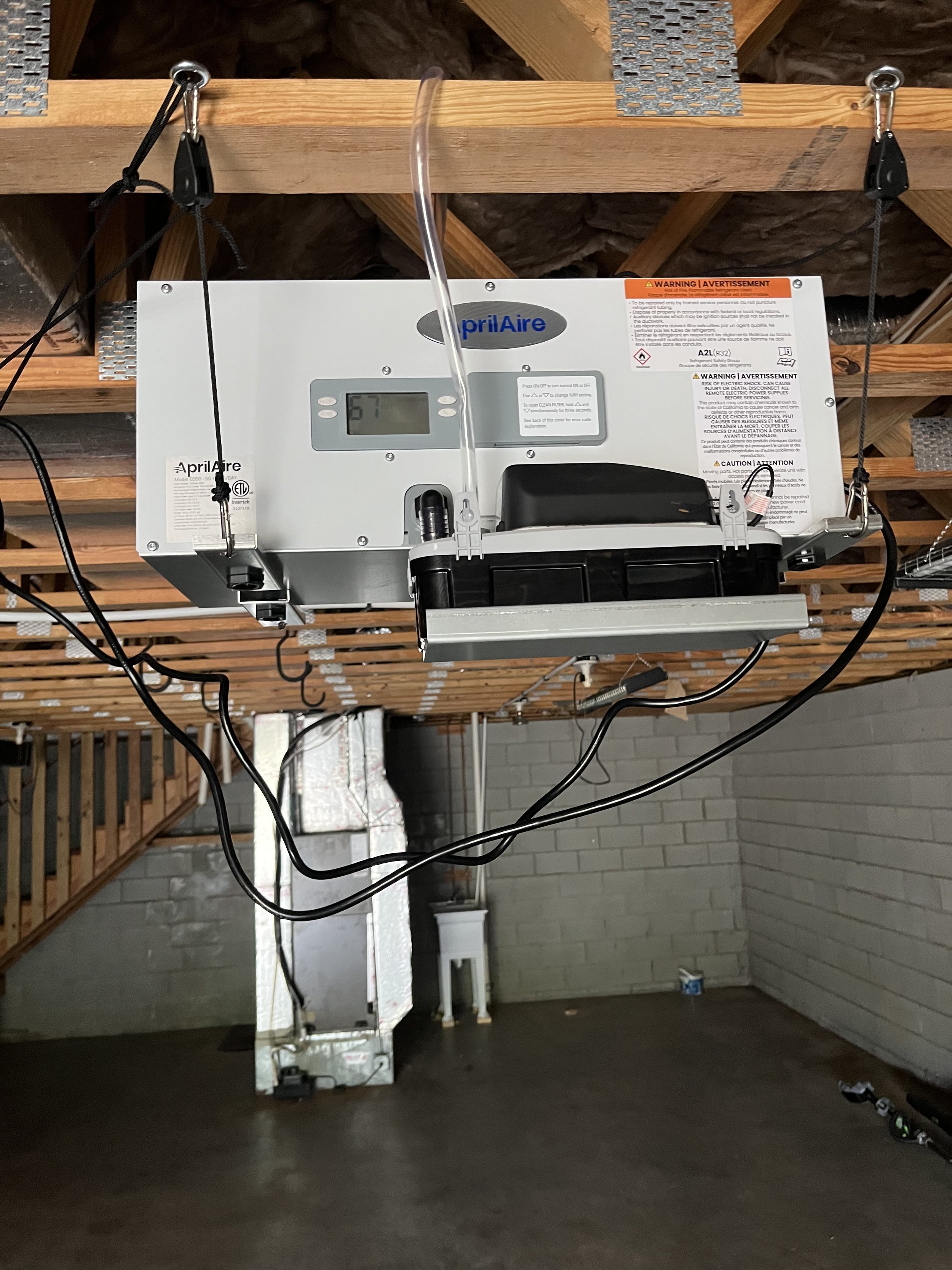 A white Aprilaire humidifier mounted on the ceiling of a basement with exposed wooden beams and insulation. There are black wires connected to the humidifier, and a black device is attached to it. The background shows a brick wall, a concrete floor, 