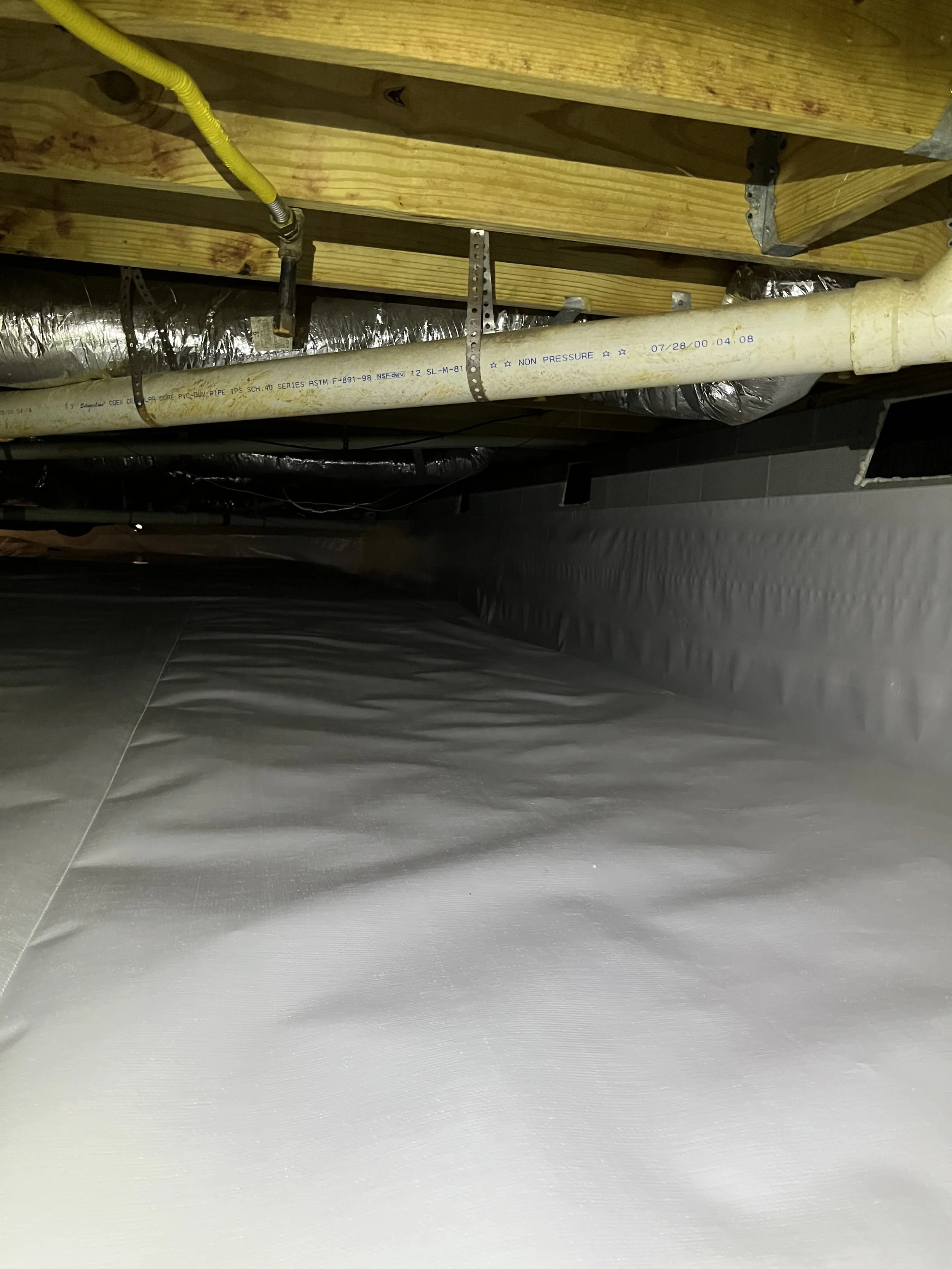 View of a basement ceiling showing wooden beams, metallic duct insulation, a white plumbing pipe, and electrical wiring.