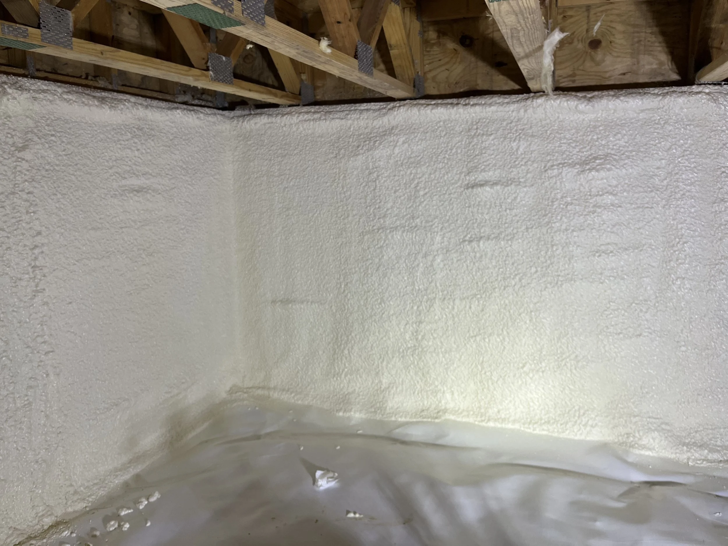 Inside view of an unfinished attic showing wooden beams and spray foam insulation on the walls.