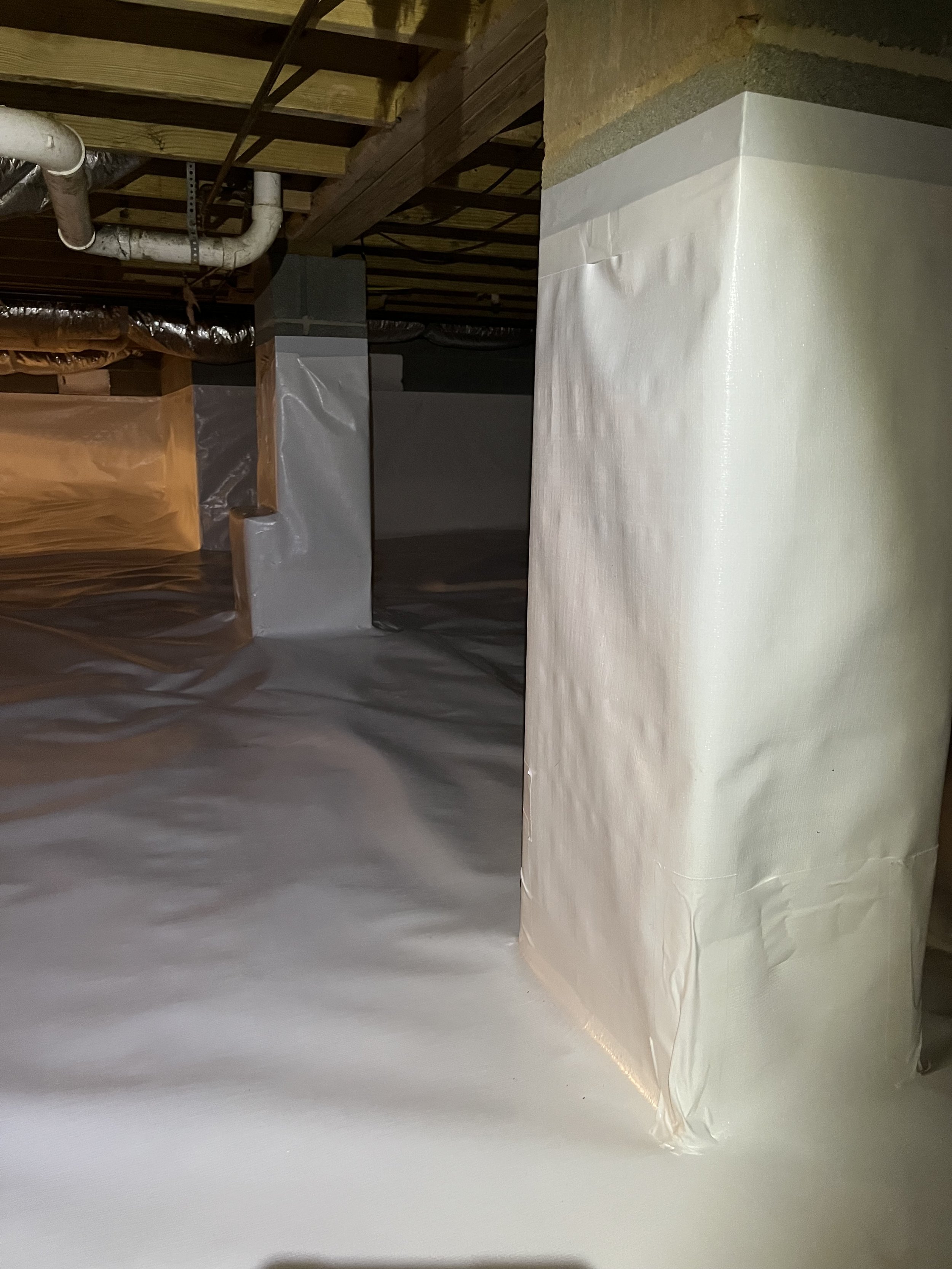 A basement construction site showing insulation, pipes, and vertical support beams. A large white wrapped object is positioned in the lower right corner.