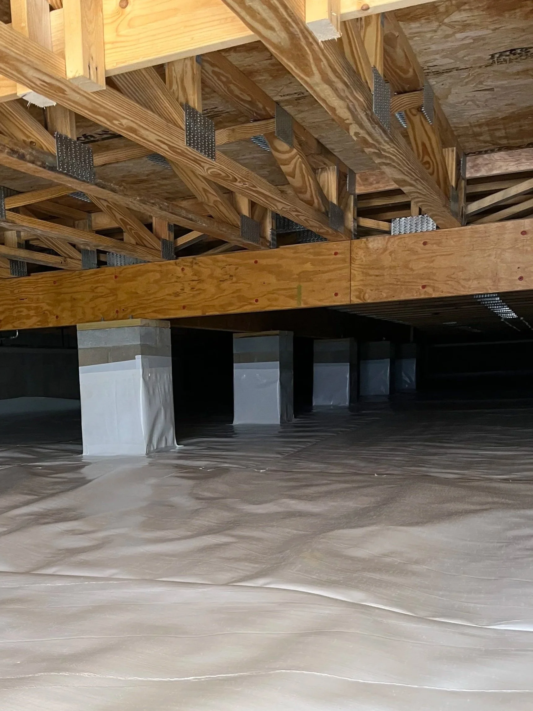 Underside of a building under construction showing wooden beams, joists, and supports with metal brackets, and a concrete foundation with plastic sheeting.