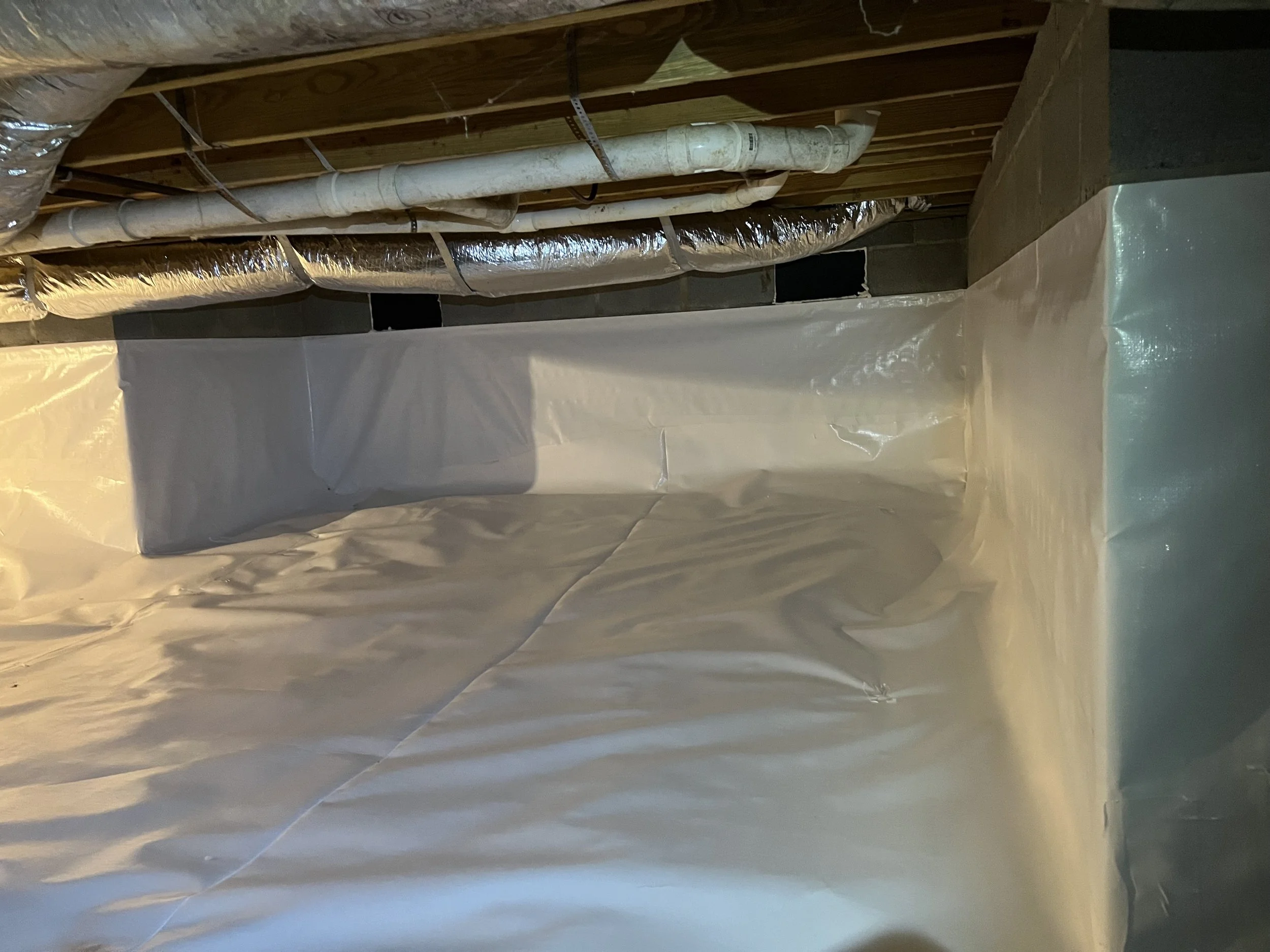 An unfinished basement ceiling with exposed wooden beams, white PVC pipes, and silver insulated ductwork, with some plastic sheeting on the walls.