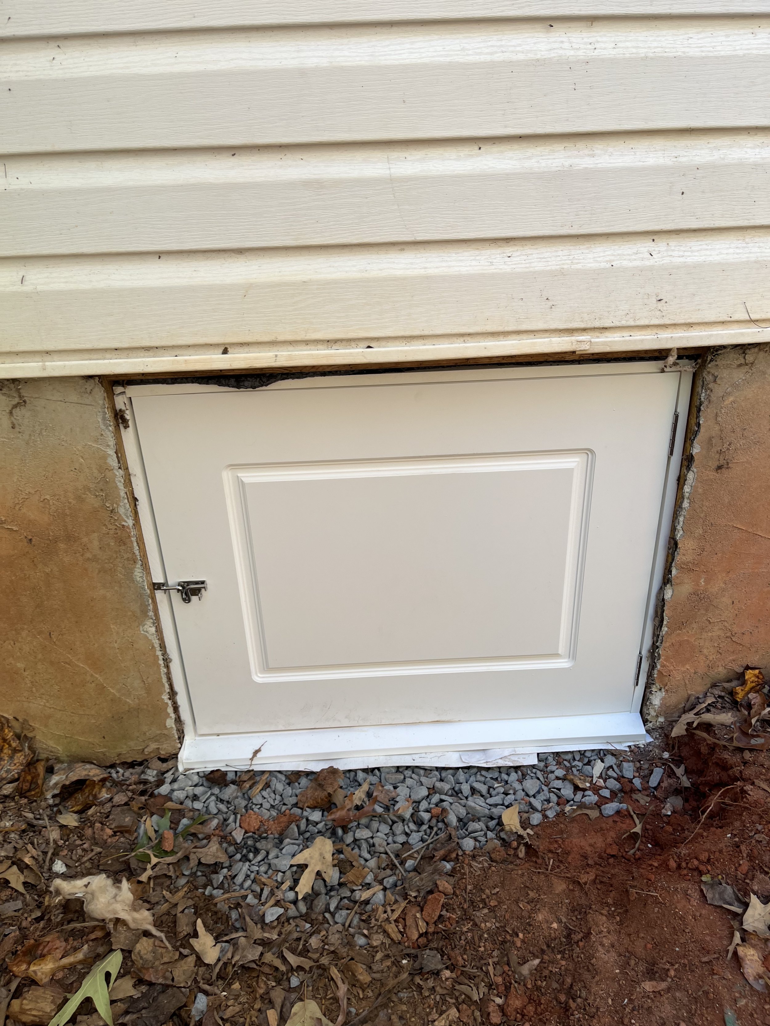 An outdoor white door with a metal latch, surrounded by exposed brick and concrete, with mulch and rocks on the ground.