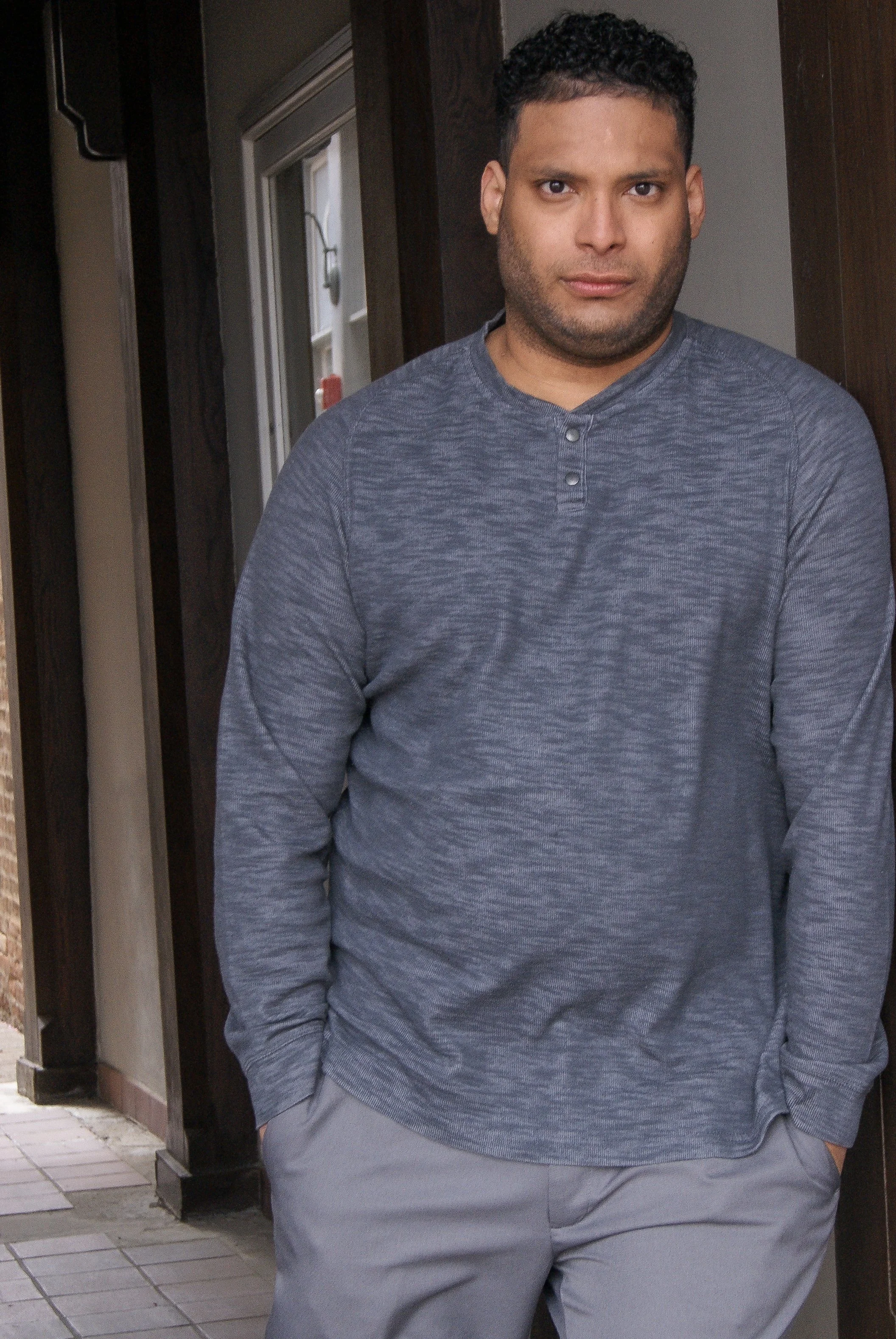 A young man with curly hair and medium skin tone wearing a long-sleeve gray shirt standing outdoors near a building with dark wooden walls and a glass door, with a tiled ground.