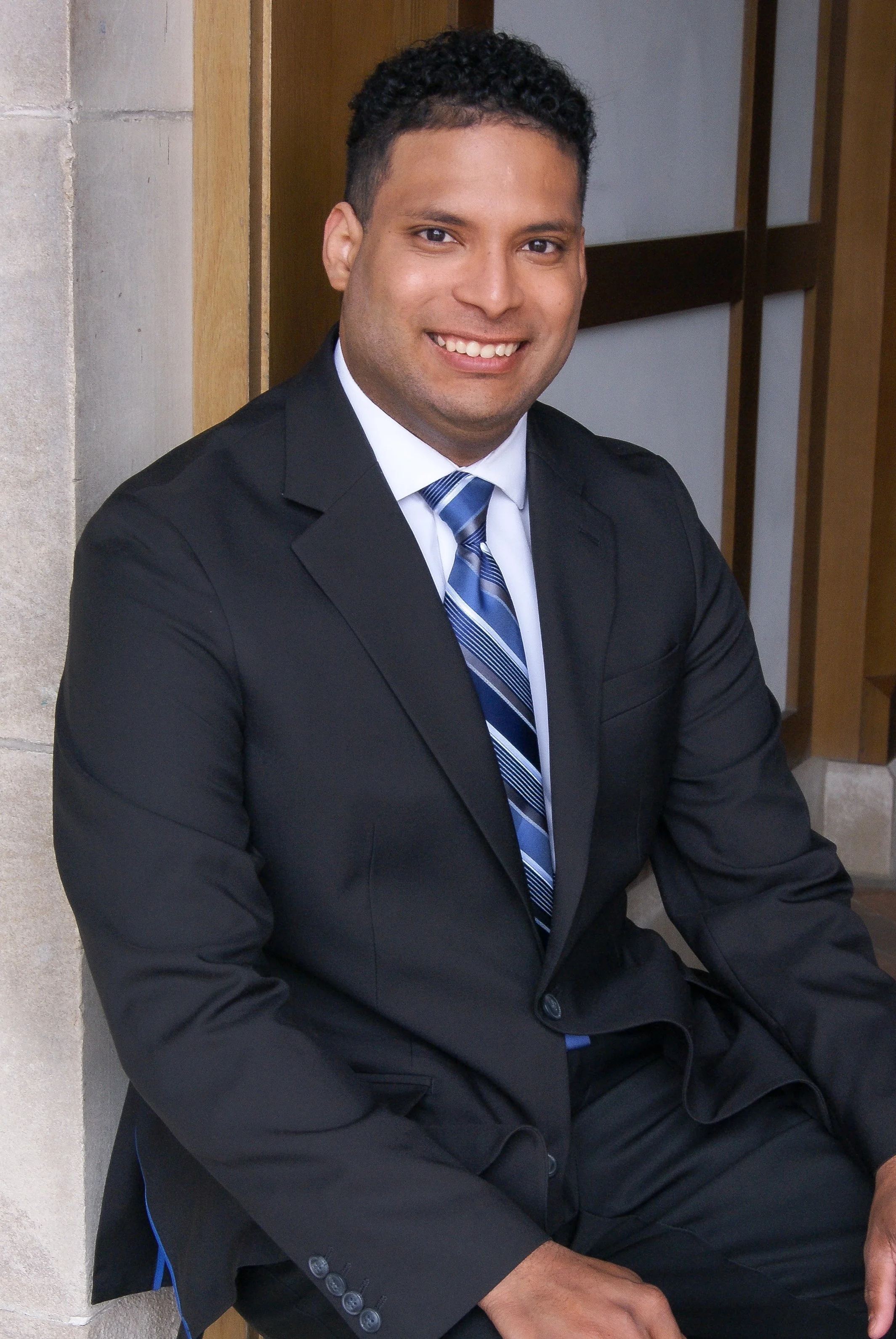 Portrait of a smiling man wearing a dark suit and patterned tie, sitting against a wooden partition.