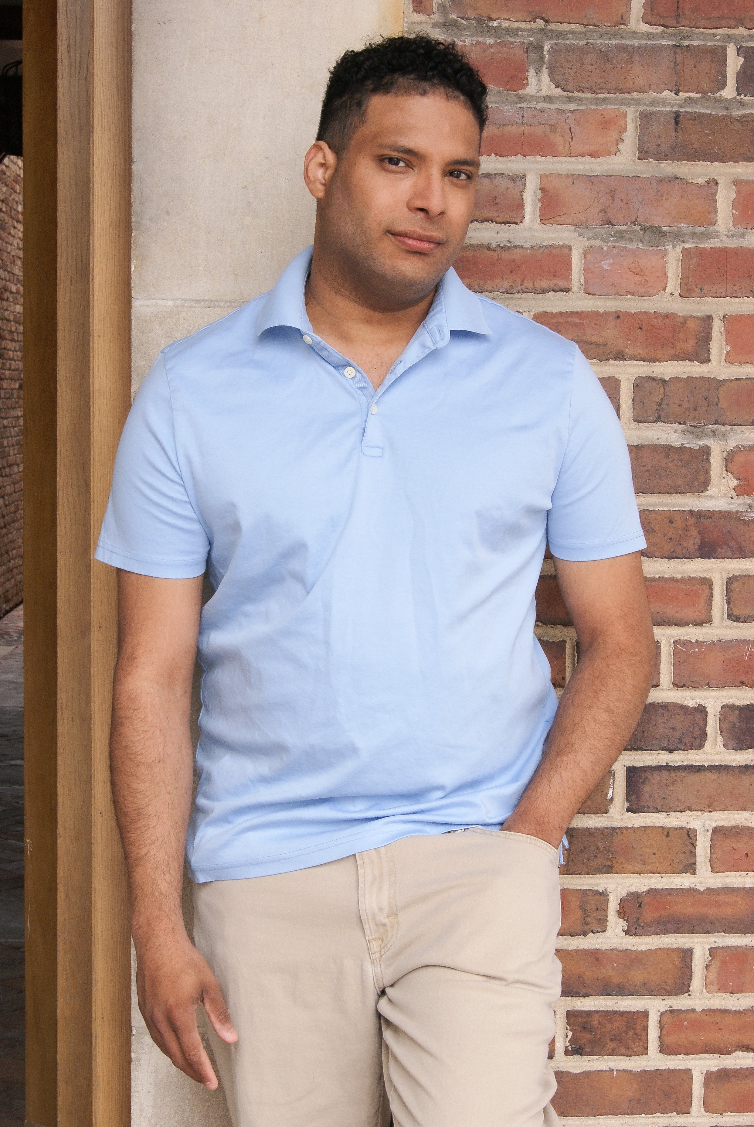 A young man standing against a brick wall, wearing a light blue short-sleeved button-up shirt and beige pants.