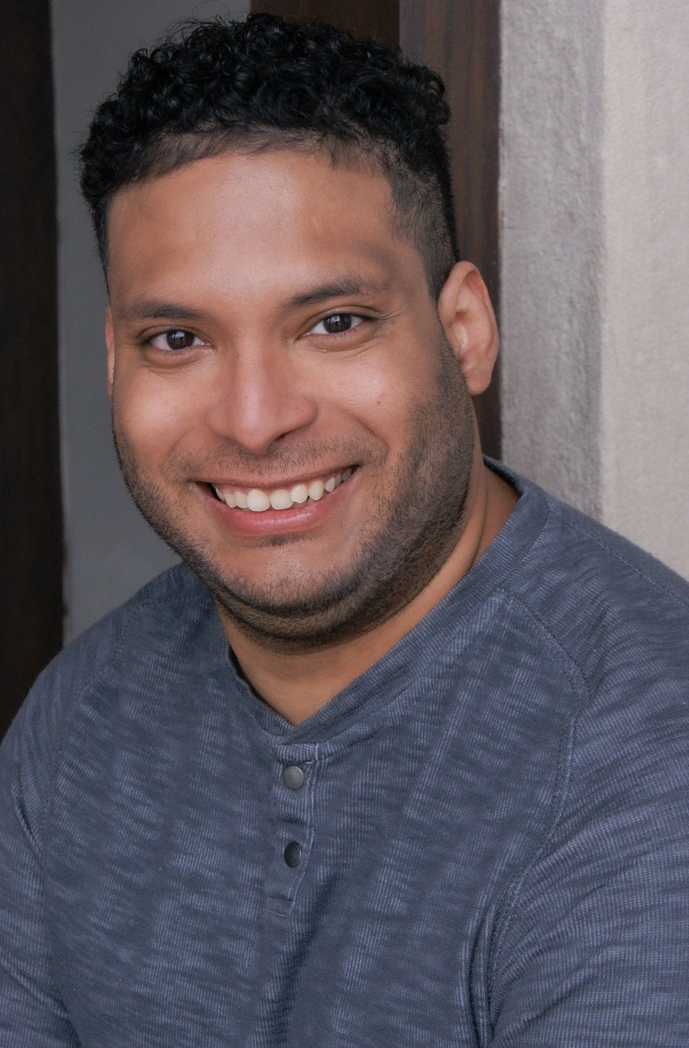 Close-up of a young man with short, curly black hair, brown eyes, and a smile showing his teeth. He is wearing a dark grey, buttoned shirt with a textured pattern. The background includes a neutral-colored wall and a wooden doorframe.