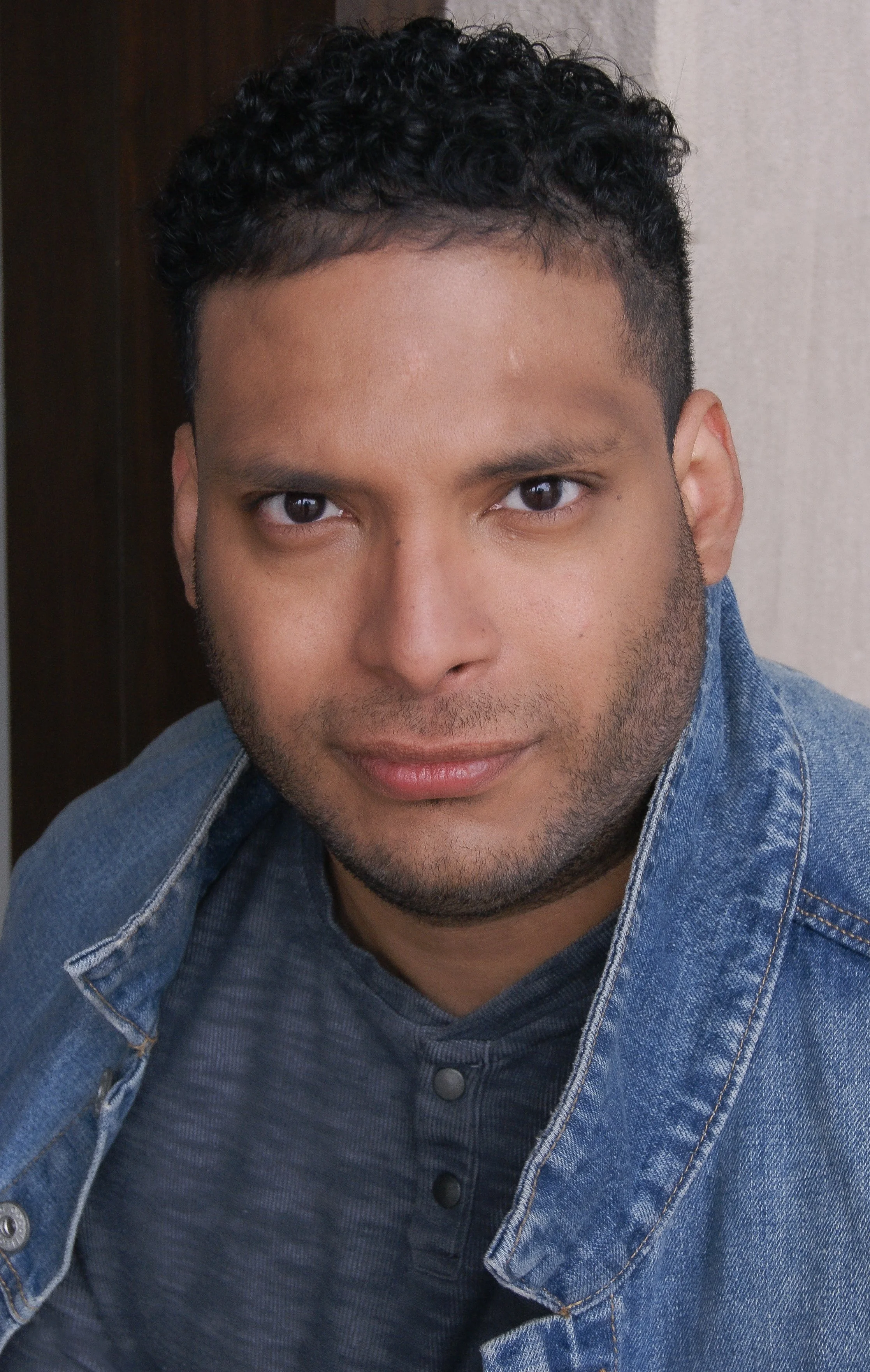 Close-up of a young man with curly black hair and brown eyes, wearing a denim jacket over a dark shirt, smiling softly at the camera.
