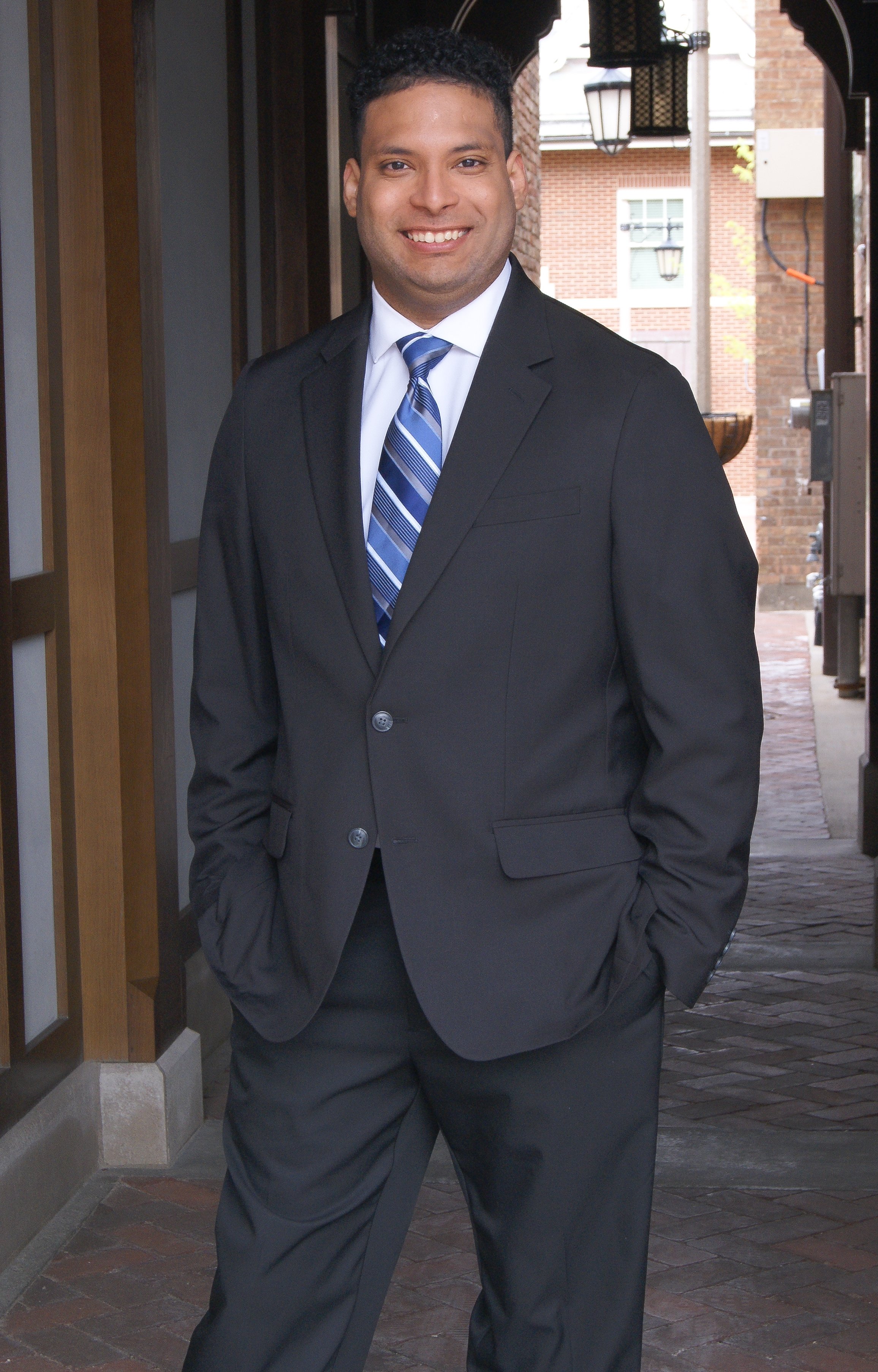 A young man in a black suit, white shirt, and striped blue tie, smiling and standing with his hands in his pockets in an outdoor setting with brick buildings and lanterns in the background.