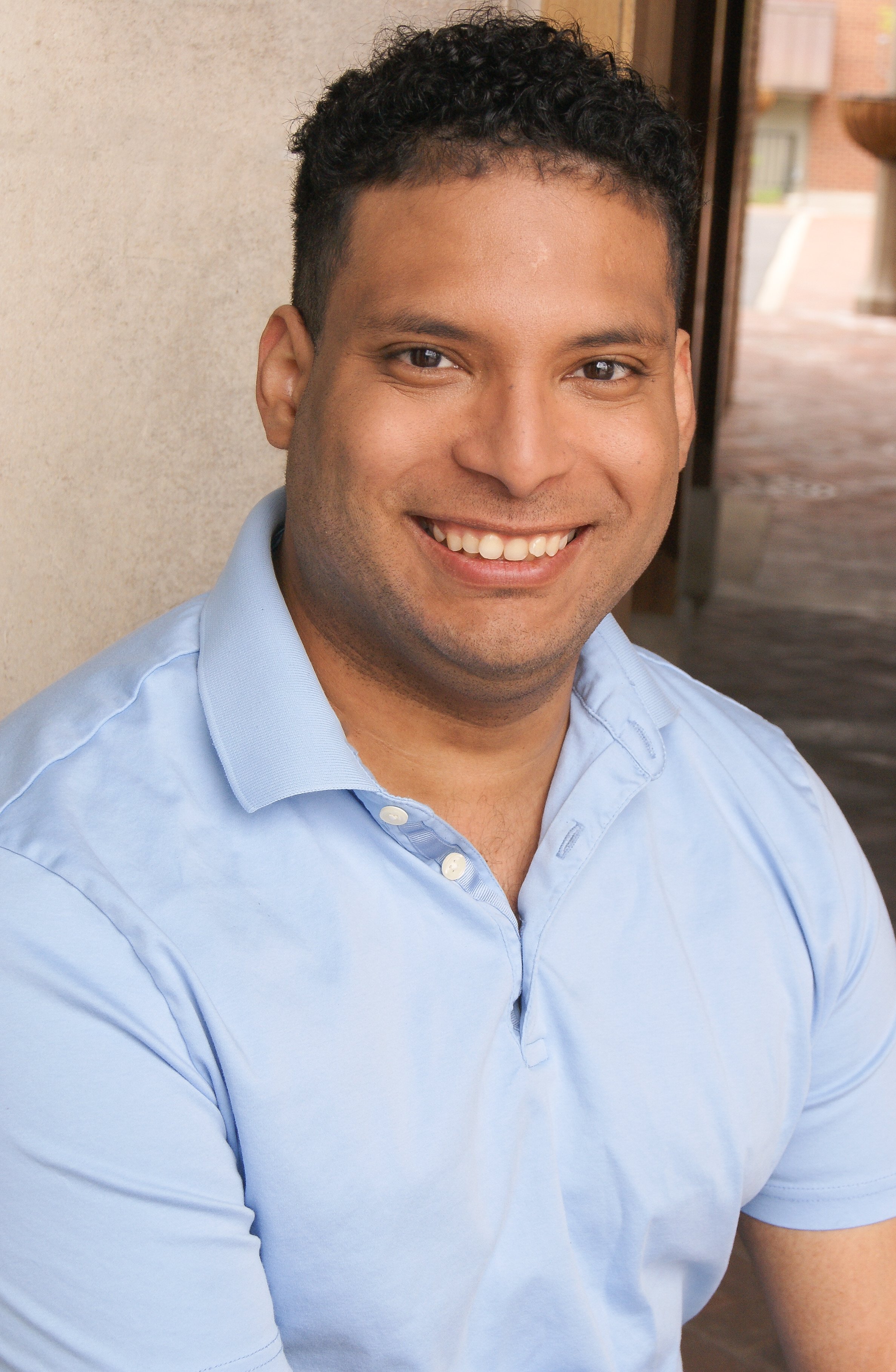 Close-up of a smiling man wearing a light blue collared shirt, standing outdoors near a beige wall.