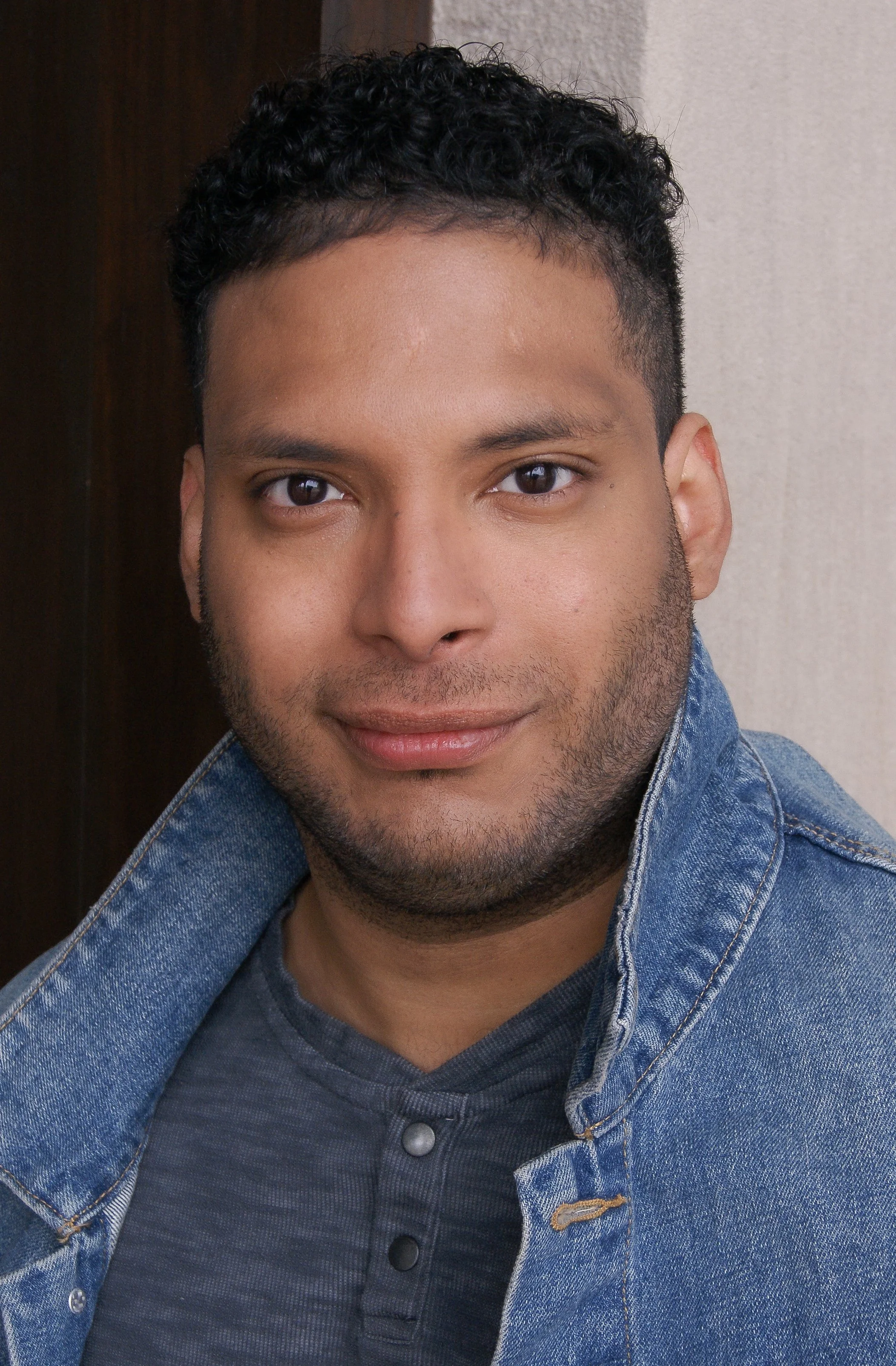 A young man with short, curly dark hair and a beard, wearing a denim jacket over a dark shirt, smiling slightly, with a neutral background.