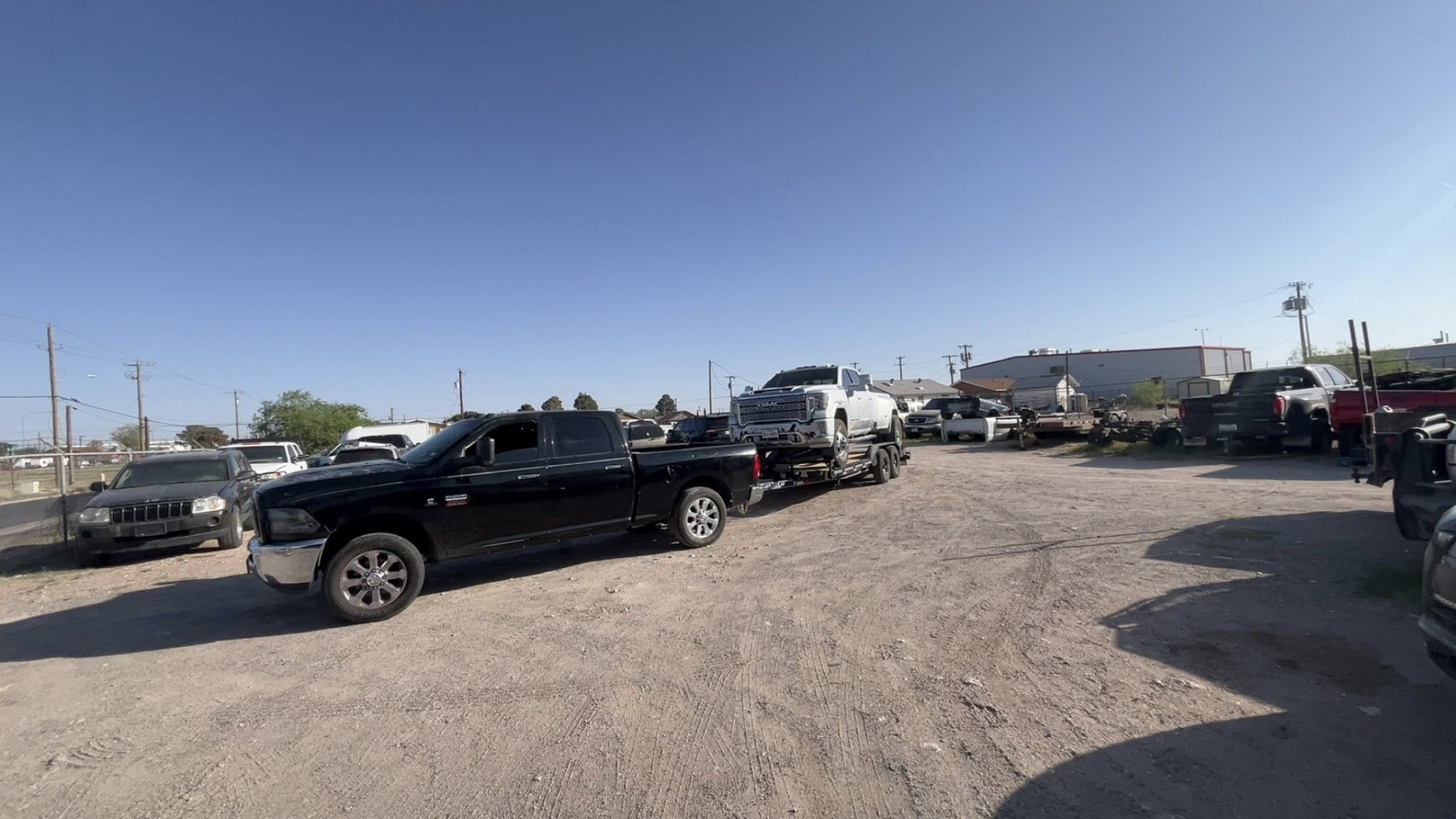 A dirt lot filled with various parked vehicles, including trucks, SUVs, and cars, with a clear blue sky overhead and utility poles in the background.