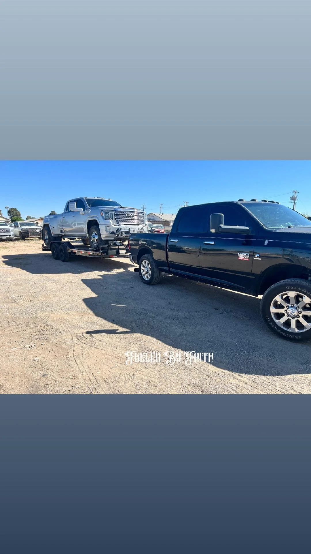 A black pickup truck towing a silver pickup truck on a flatbed trailer in a car lot on a clear, sunny day.
