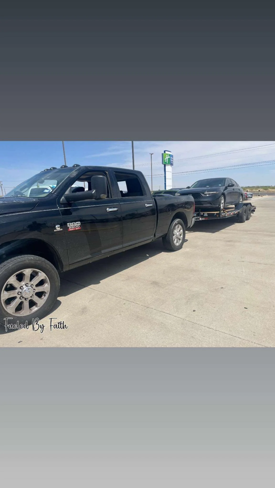 Black pickup truck transporting a car on a flatbed trailer at a parking lot with a Holiday Inn sign in the background.