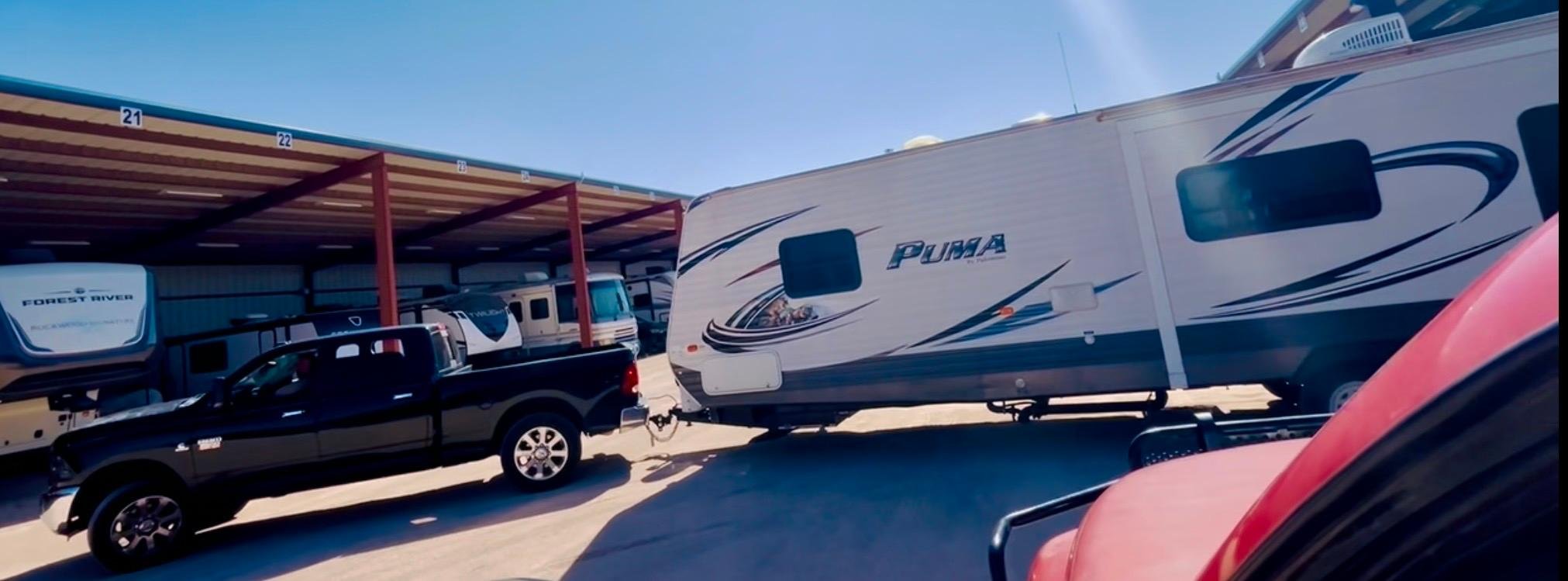 A black pickup truck towing a large white Puma RV trailer parked at a storage facility with multiple other RVs and trailers under a mostly clear blue sky.
