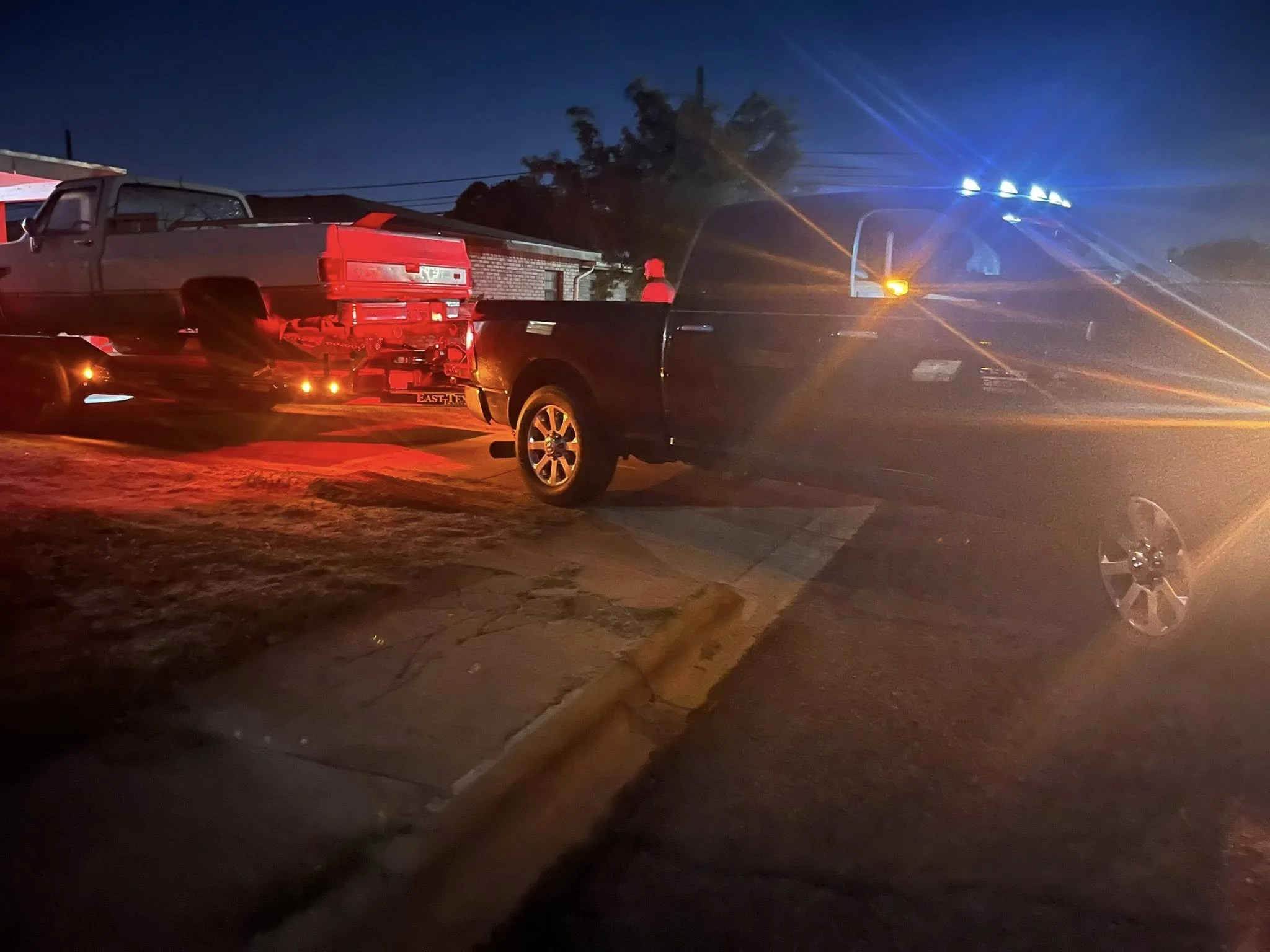 Nighttime scene with a black pickup truck, a flatbed trailer carrying a red vehicle, and a person wearing a red hat standing in front of a building. Emergency vehicle with blue flashing lights is present.