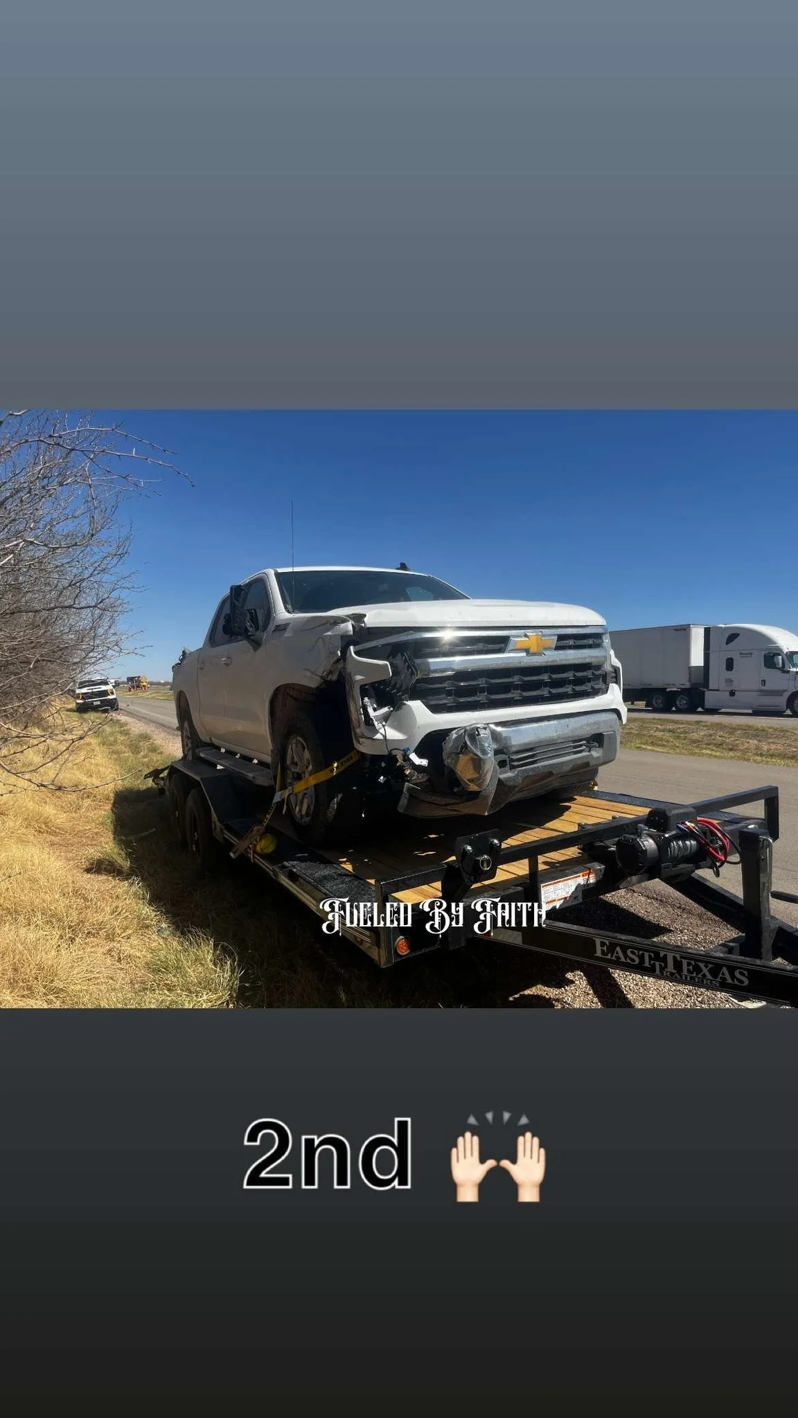 A damaged gray Chevrolet pickup truck on a flatbed trailer on the side of a highway. The front of the truck is visibly wrecked. The sky is clear and blue, with other trucks passing by in the background. Text on the image says 'FUELED BY FAITH' and '2