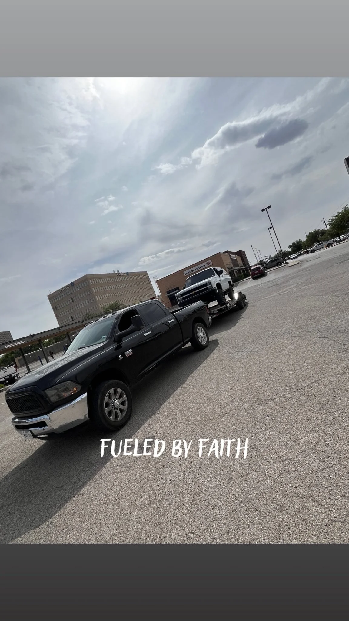 A black pickup truck towing a smaller white vehicle in a parking lot with several buildings and cars, cloudy sky overhead, and text overlaid that reads 'FUELED BY FAITH'.