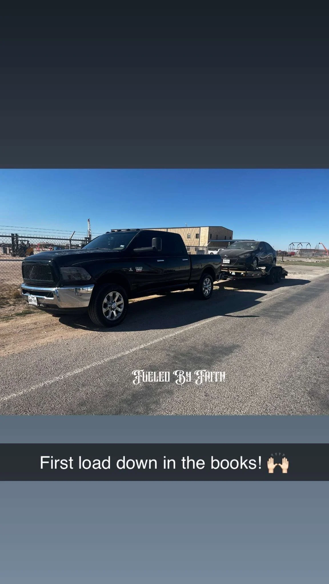 A black pickup truck is parked on the side of a road, pulling a trailer with a black car loaded on it. The scene is outdoors under a clear blue sky with some industrial buildings and construction equipment visible in the background. Text overlay read