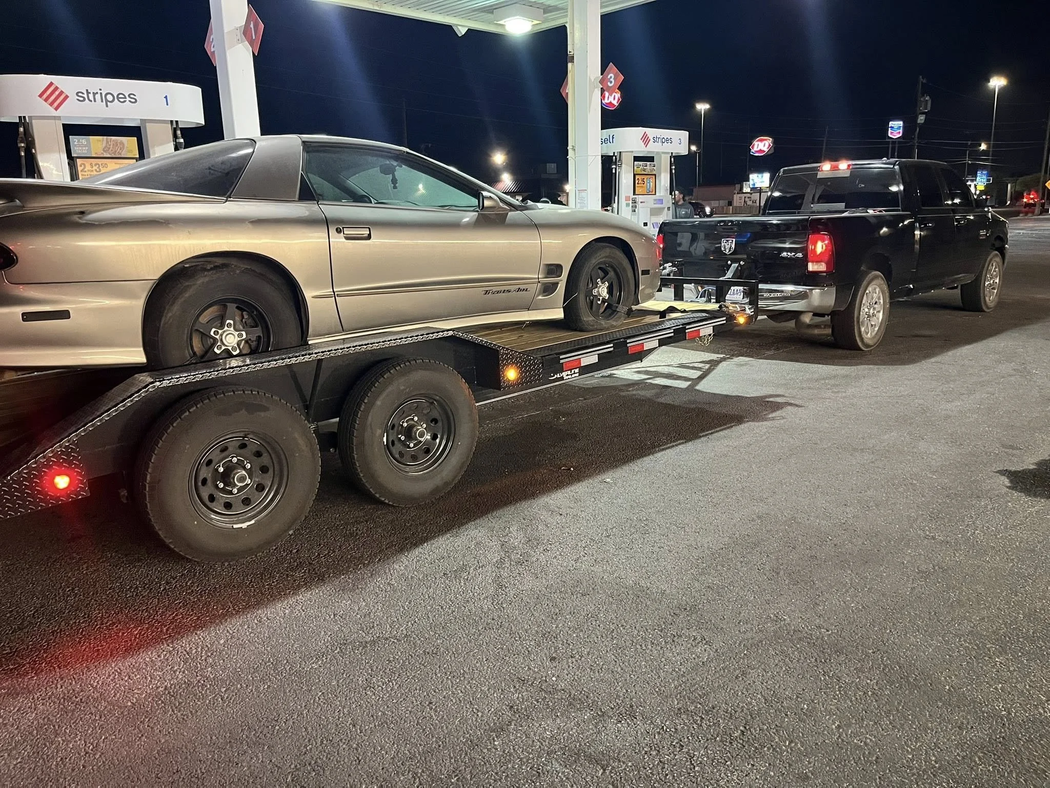 A black pickup truck on a flatbed trailer at a gas station, with the trailer hooked to a silver sports car.
