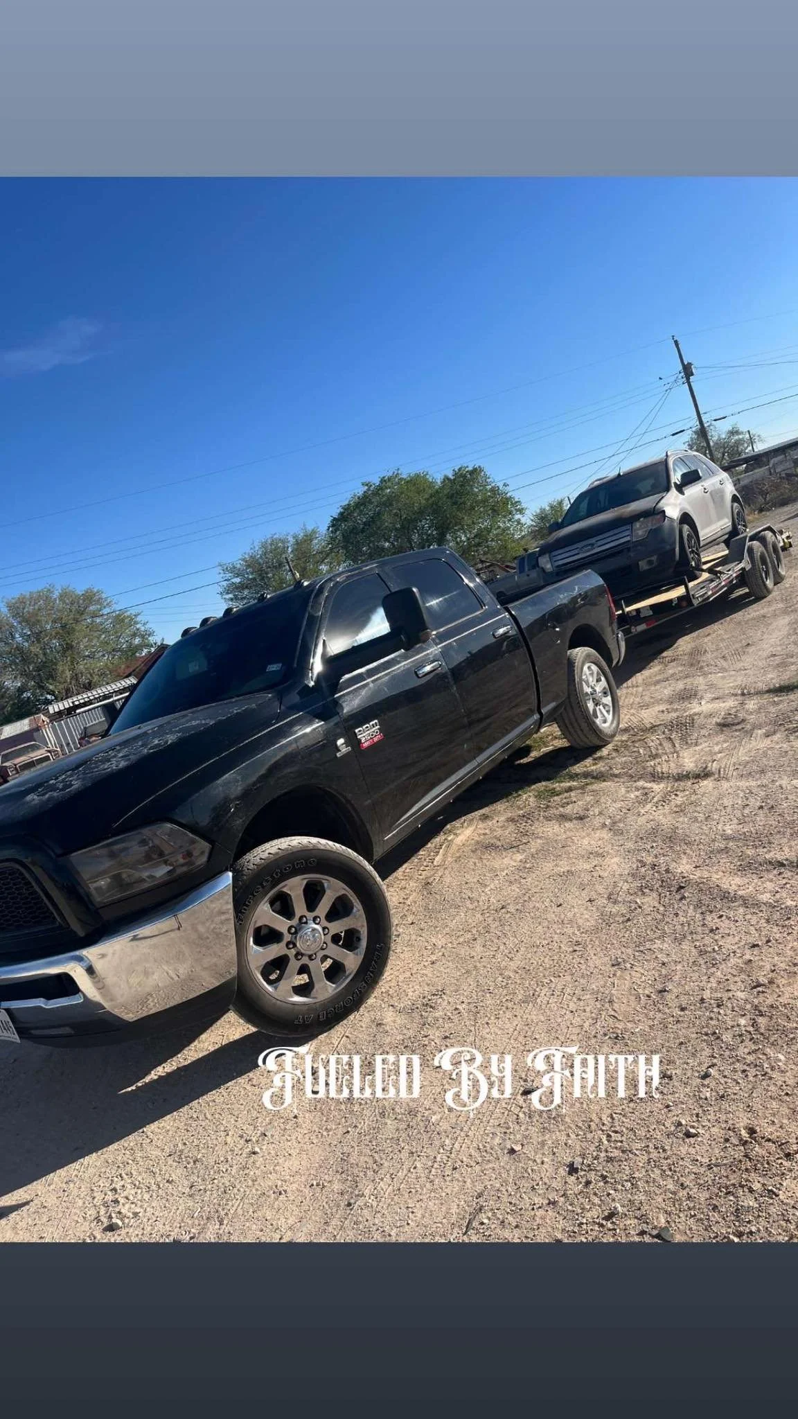 A black pickup truck towing a silver SUV on a trailer in a dirt lot with trees and utility poles in the background.