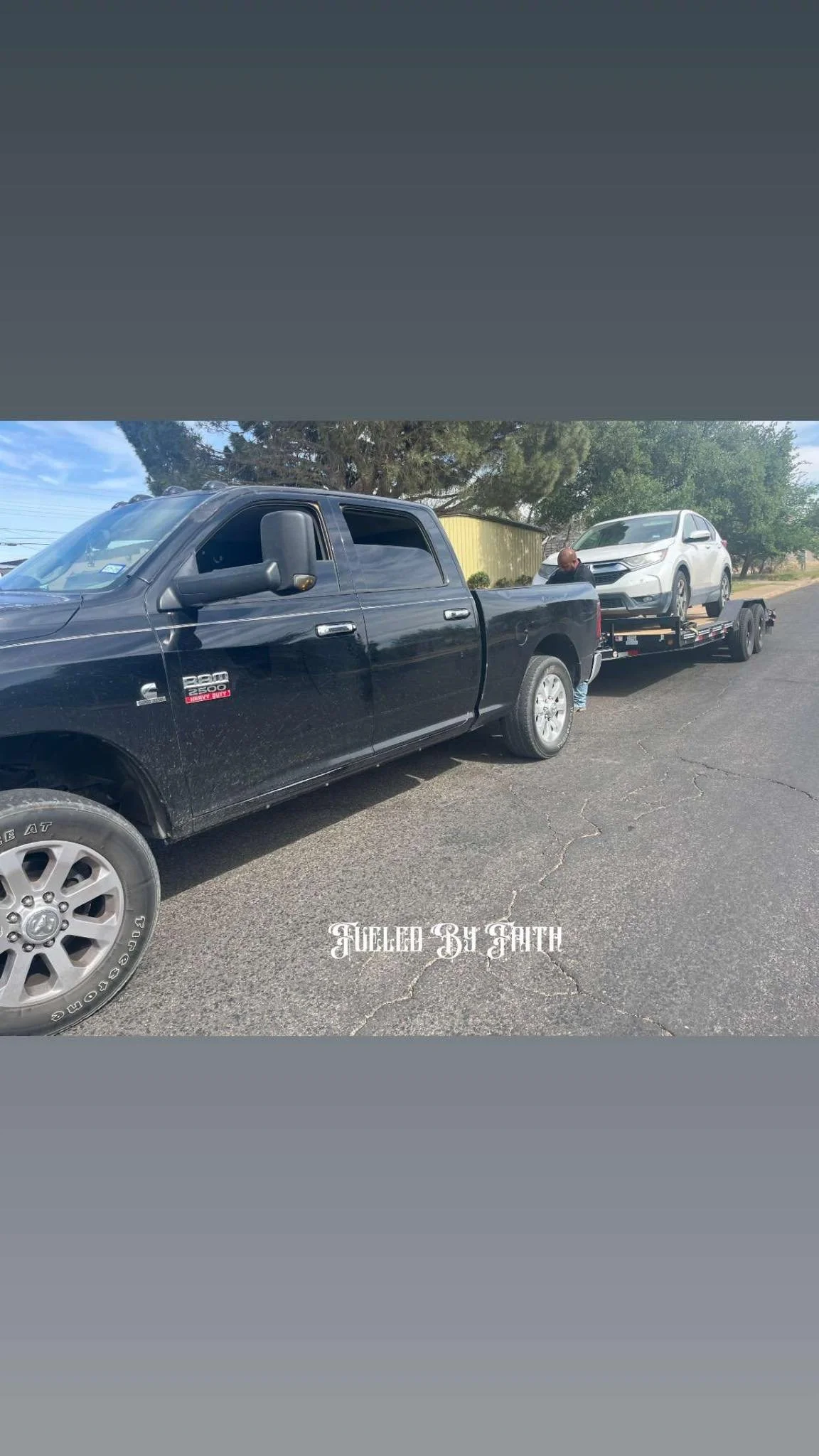 A black pickup truck with a partially open window, towing a white SUV on a flatbed trailer on a paved road, with trees and a yellow building in the background, and the text "FILLED BY FAITH" at the bottom.